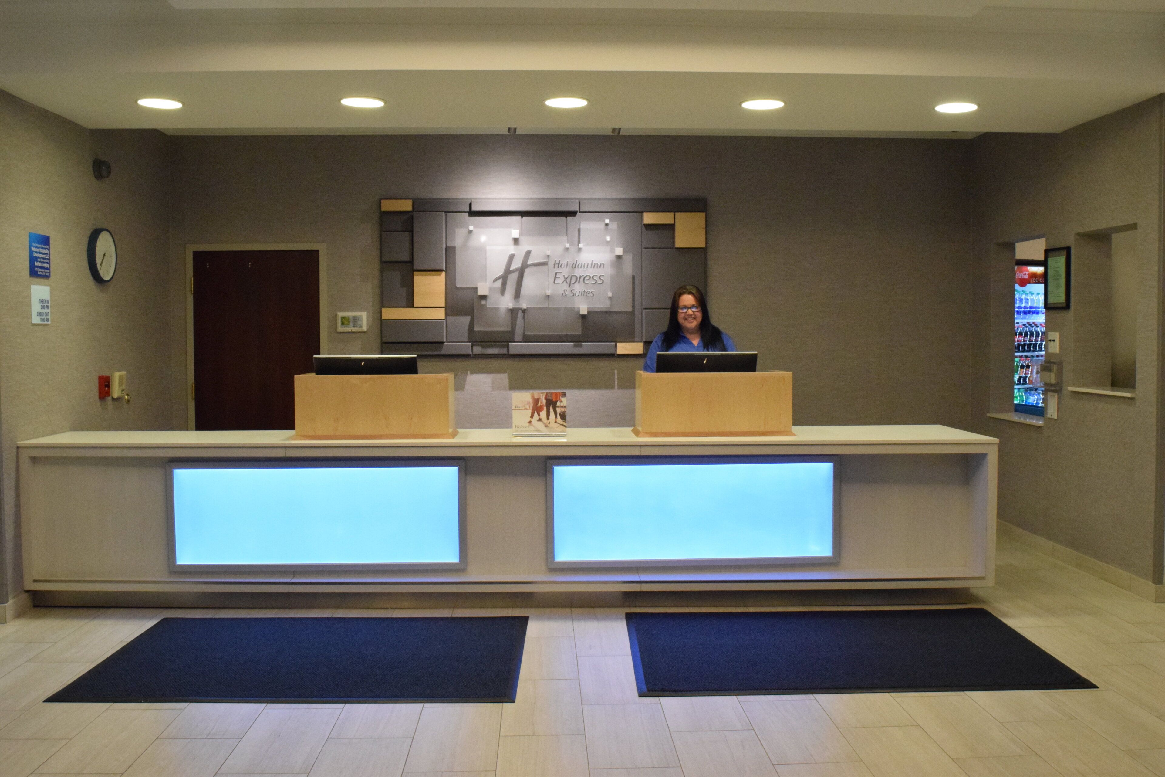 A hotel reception desk with a digital display, a clock, and a Coca-Cola refrigerator.