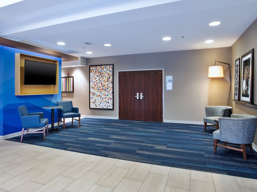 A hotel lobby with a blue accent wall, a flat-screen TV, a large abstract painting, a wooden door, and two seating areas with chairs.