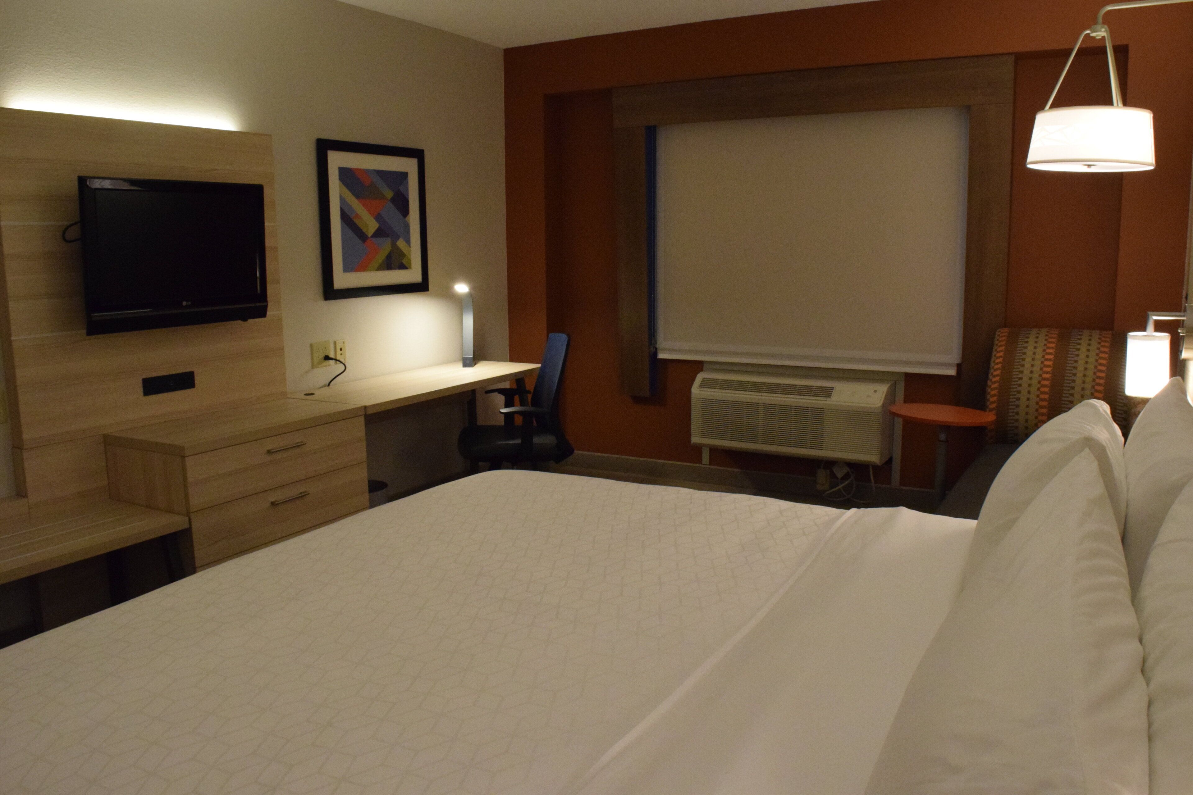 Habitación de hotel con una cama, un escritorio con lámpara, televisión, ventana con persiana y radiador.