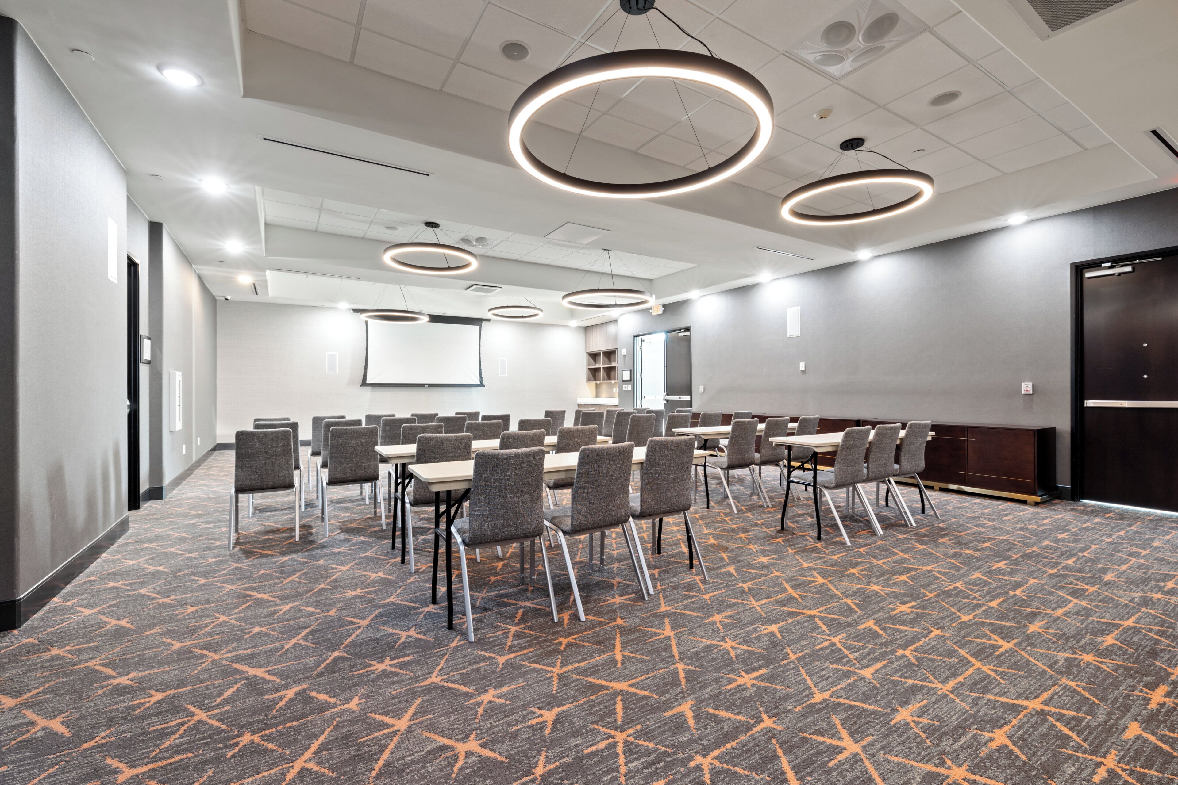 A conference room with a carpeted floor, multiple tables with chairs, and overhead circular lighting fixtures.