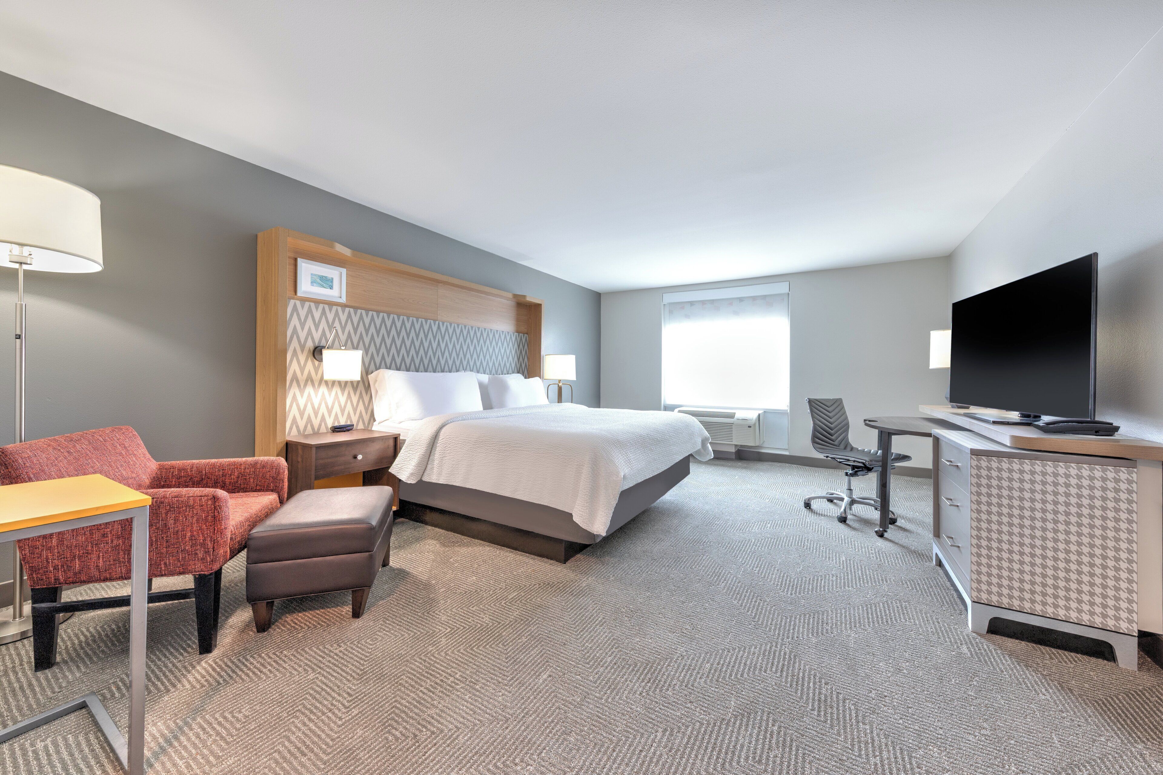 A hotel room with a bed, a desk, a chair, a TV, and a lamp.