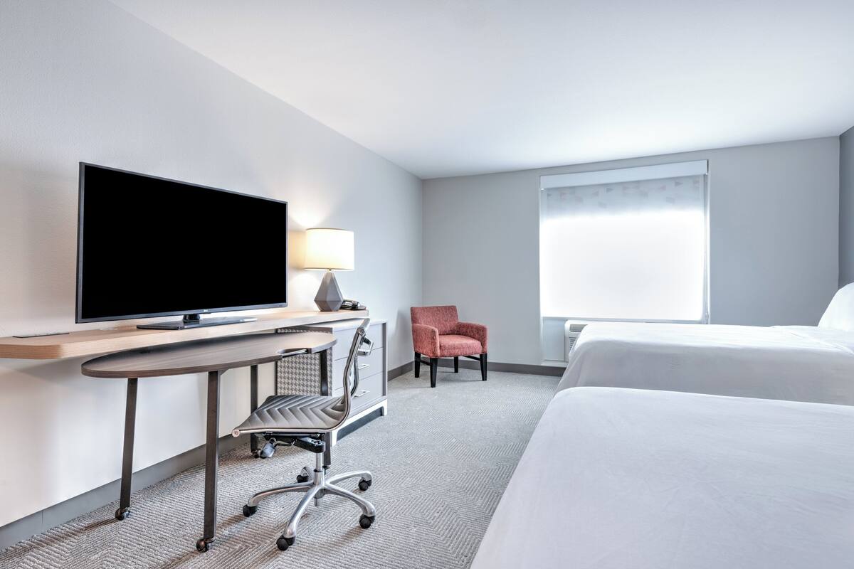 Ein Hotelzimmer mit Flachbildfernseher, Schreibtisch mit Lampe, Stuhl und zwei Betten.