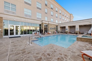 A hotel pool area with a stone-tiled deck, lounge chairs, and a building with large windows and a glass entrance.