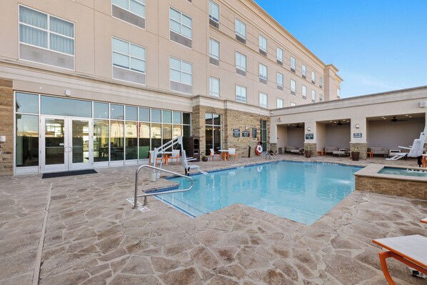 A hotel pool area with a stone-tiled deck, lounge chairs, and a building with large windows and a glass entrance. - Holiday Inn Killeen – Fort Hood Area by IHG (Killeen)