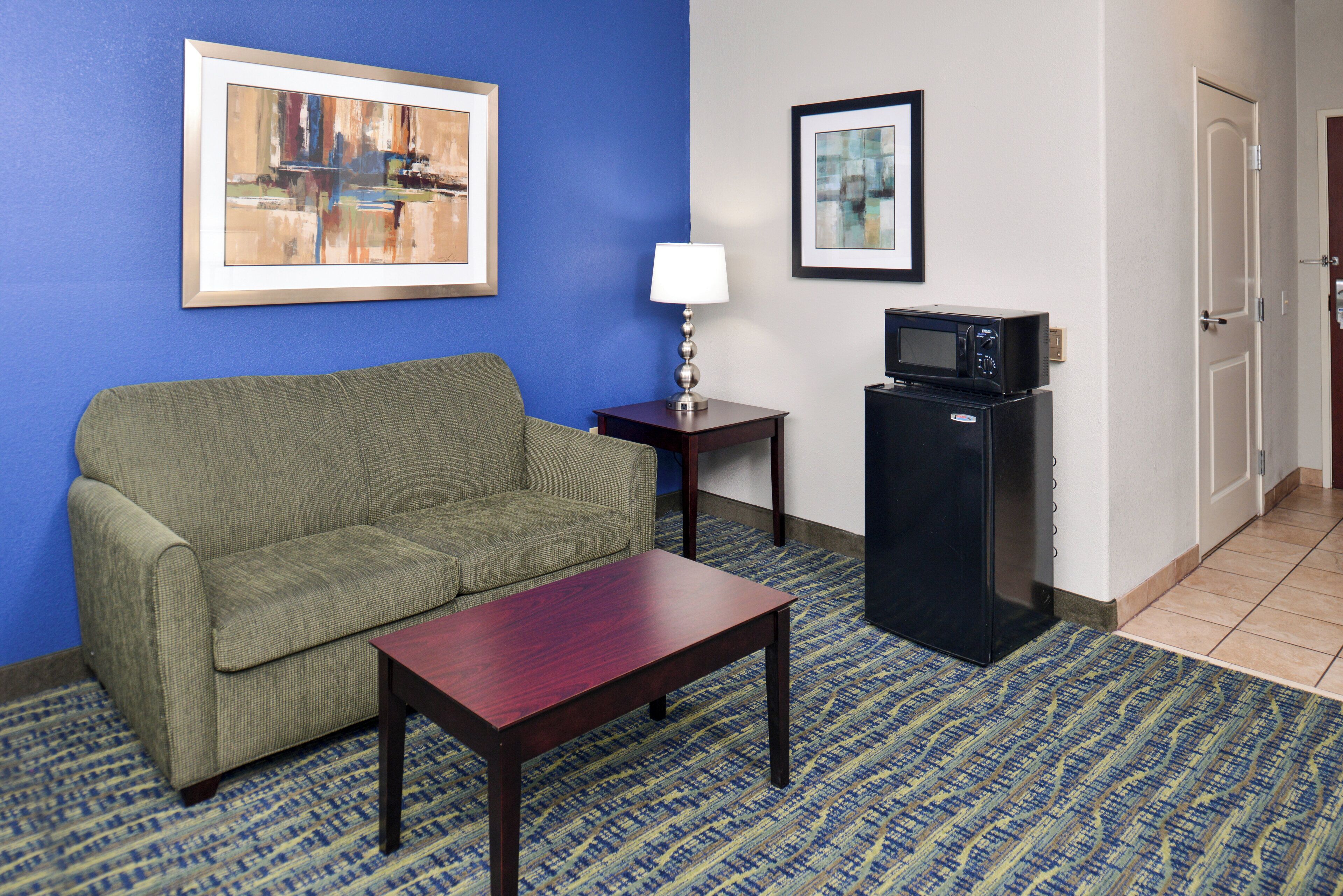 A hotel room with a green sofa, a wooden table, a lamp, a microwave, and two framed pictures on the wall.