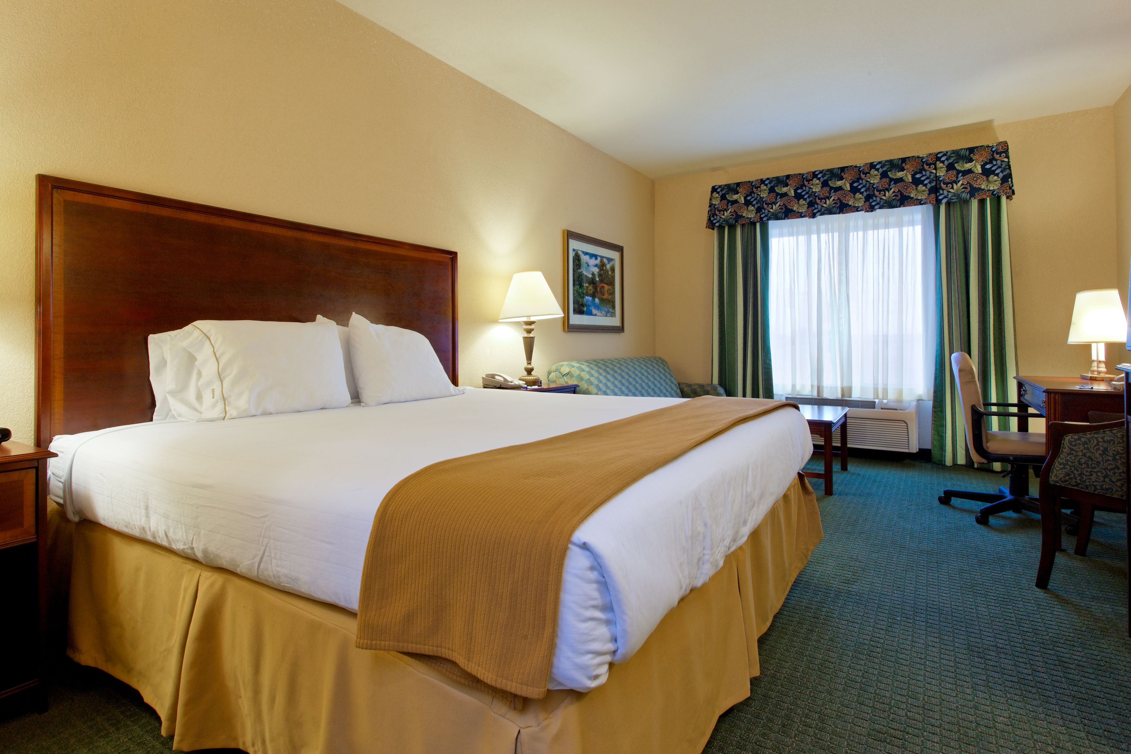 A hotel room with a large bed, a desk with a chair, a window with curtains, and a painting on the wall.