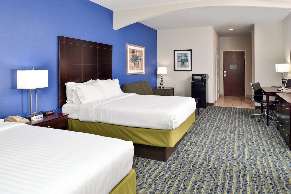 Quarto de hotel com duas camas, uma escrivaninha, uma cadeira, um abajur e uma porta.