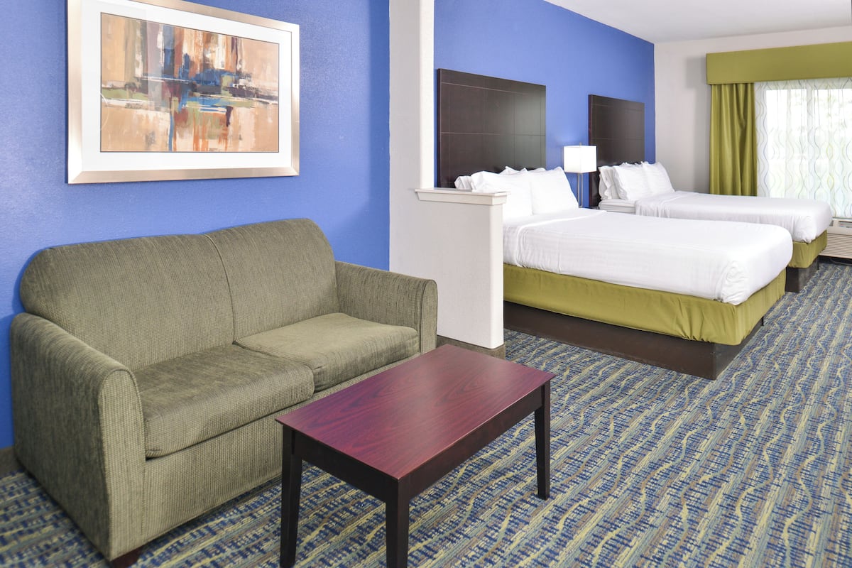 Ein Hotelzimmer mit einem grünen Sofa, einem Holztisch, zwei Betten mit weißer Bettwäsche, einem Bild an der Wand und einem gemusterten Teppich.