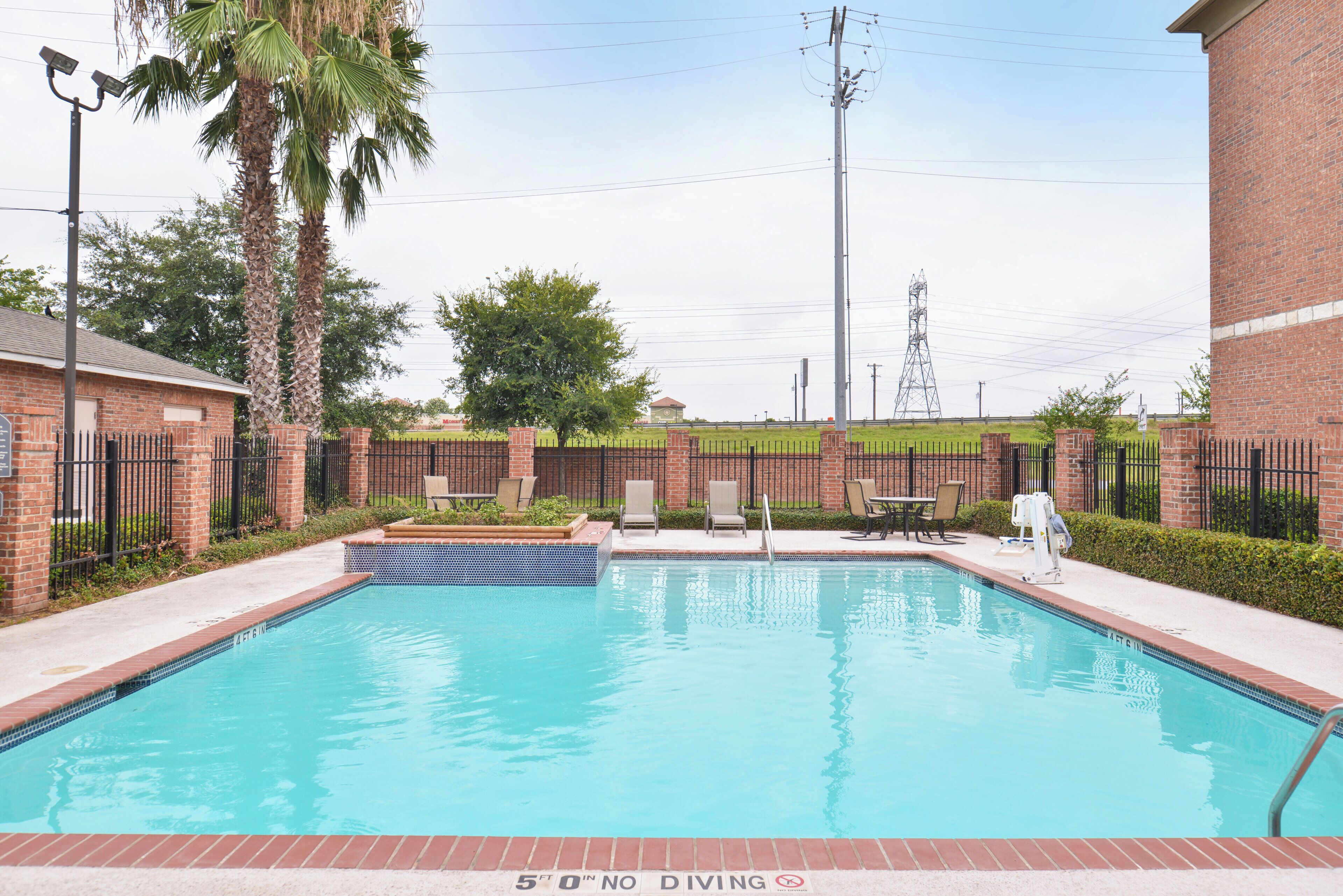 Una piscina rectangular con borde de azulejos, rodeada por un muro de ladrillo y una valla metálica.