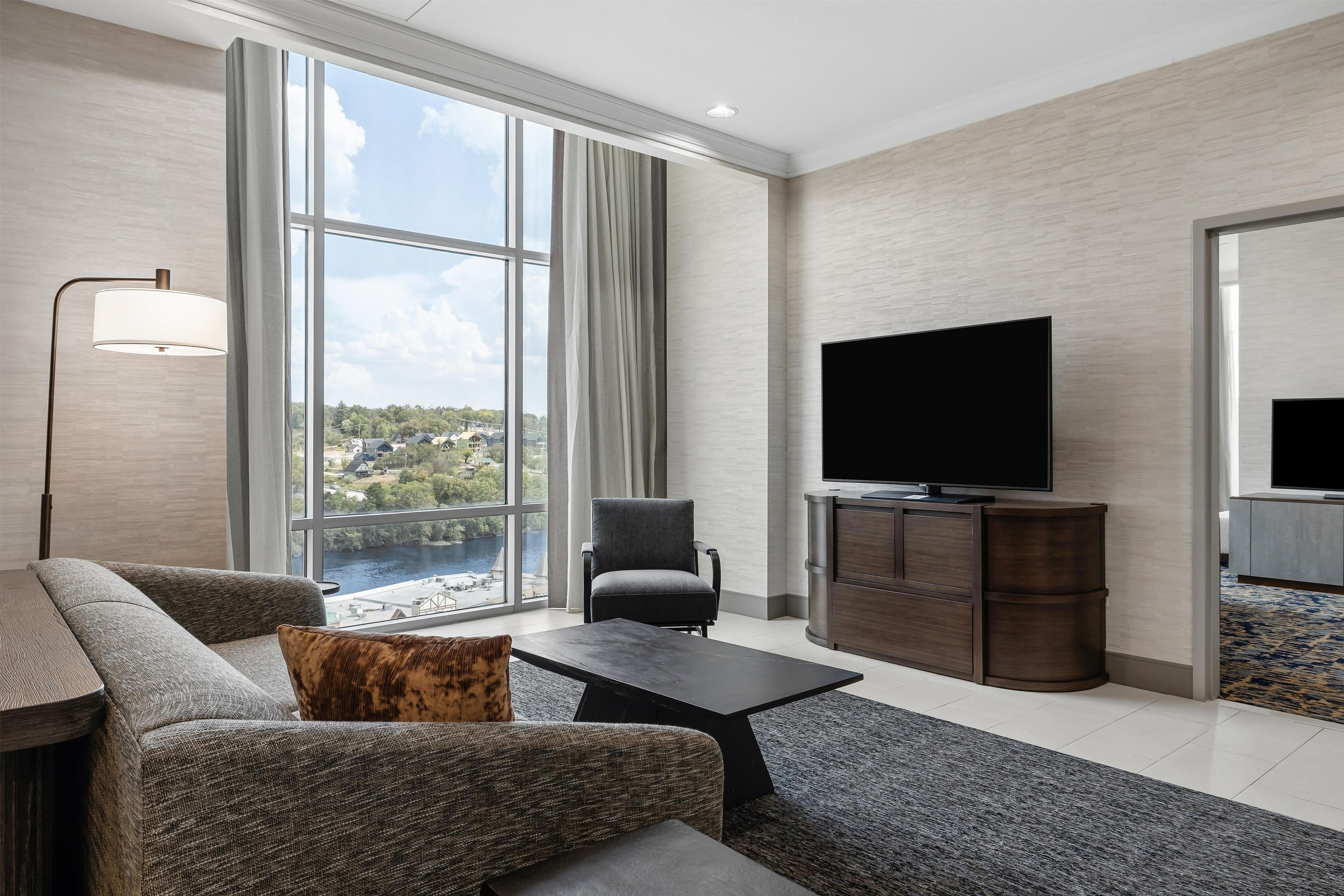 Una habitación de hotel moderna con un ventanal grande, un televisor de pantalla plana, un sofá, una silla y una pequeña mesa.