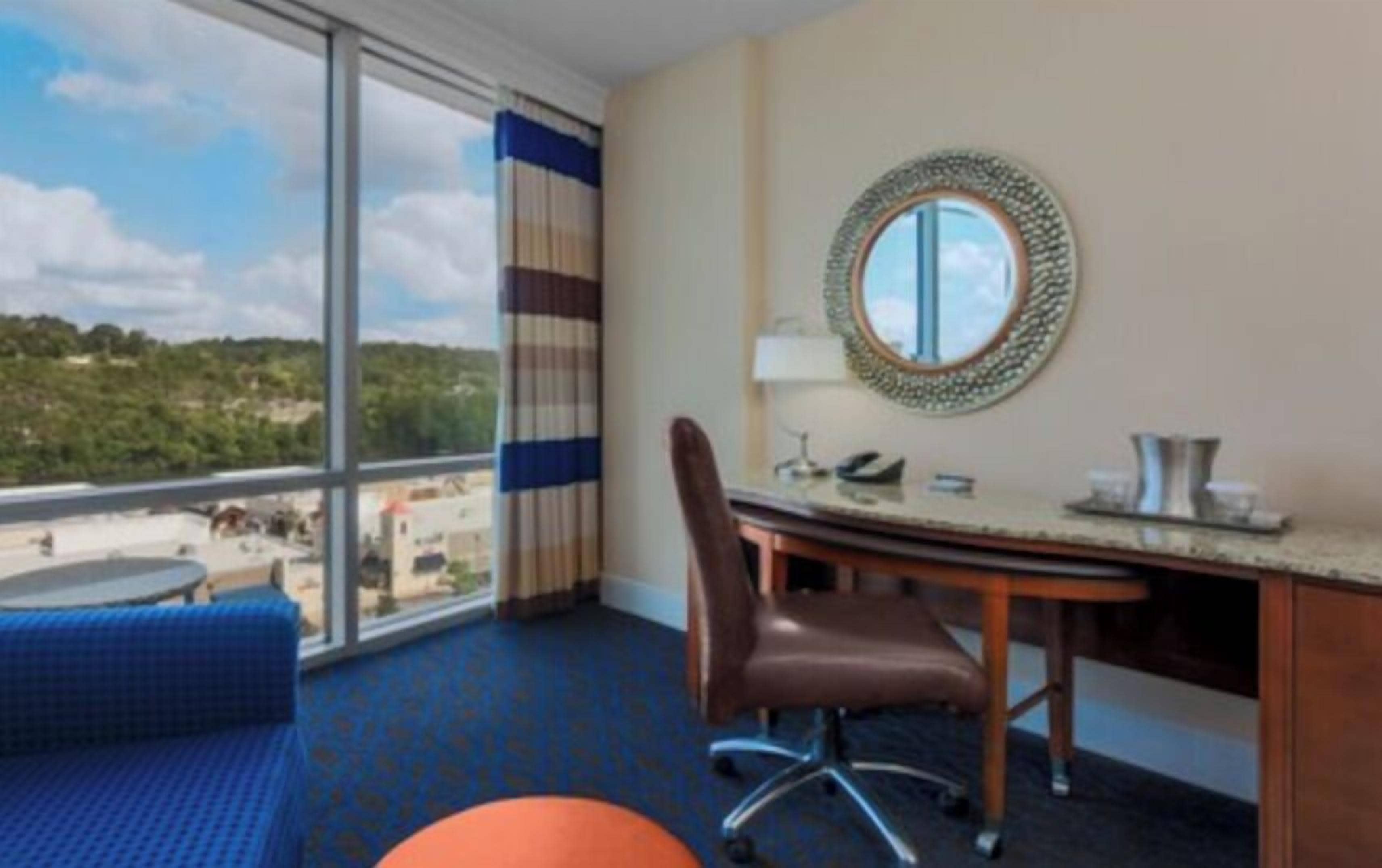 Een hotelkamer met een groot raam, een bureau met stoel, een ronde spiegel en een blauw vloerkleed.