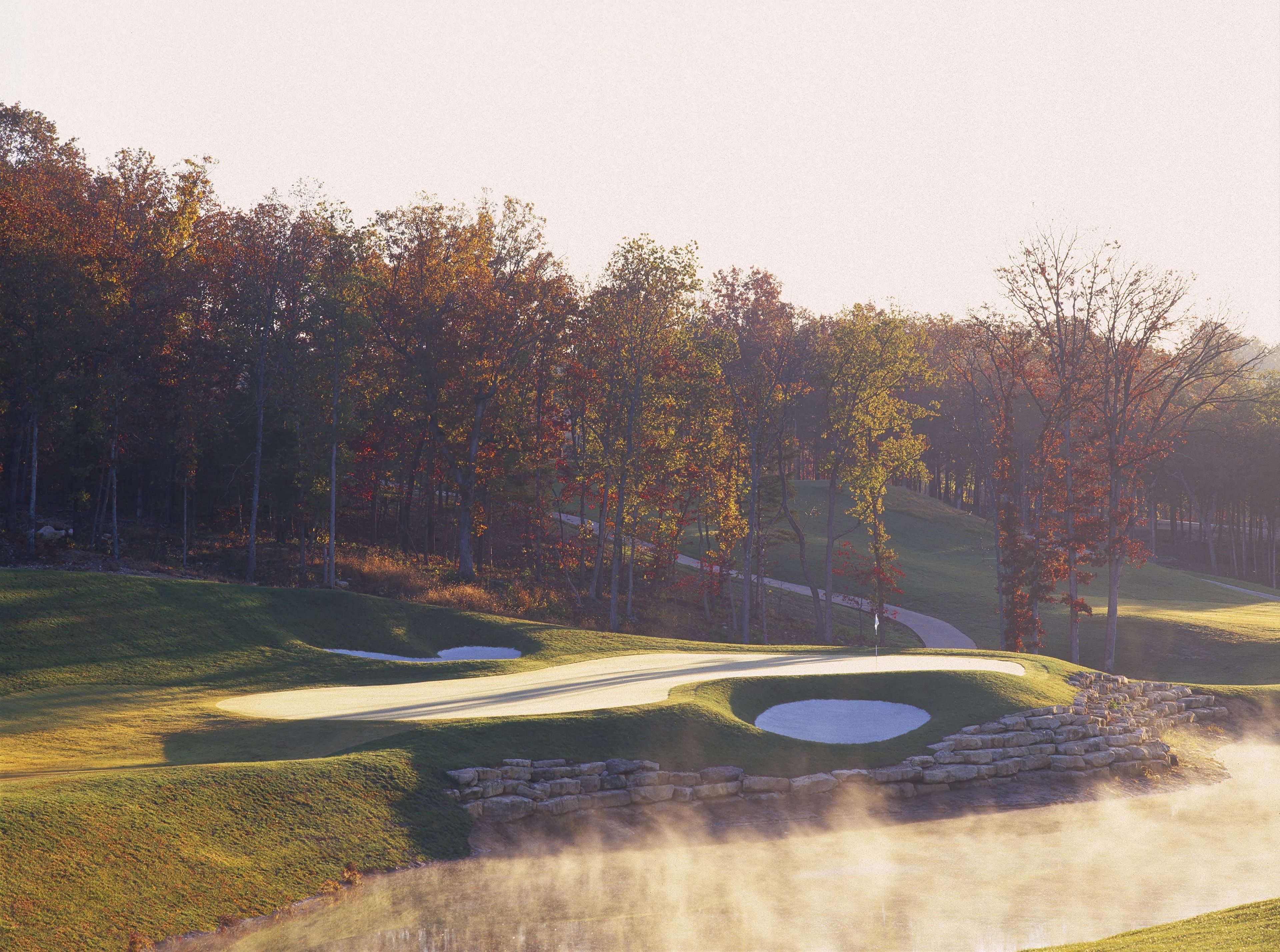 Un campo de golf con un fairway bien cuidado, búnkeres de arena y una zona boscosa con follaje otoñal.