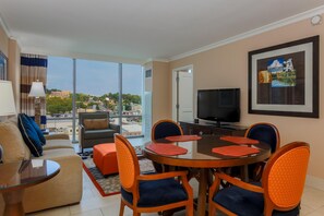 Habitación de hotel con zona de comedor, sofá, televisión y un amplio ventanal con vistas a la ciudad.