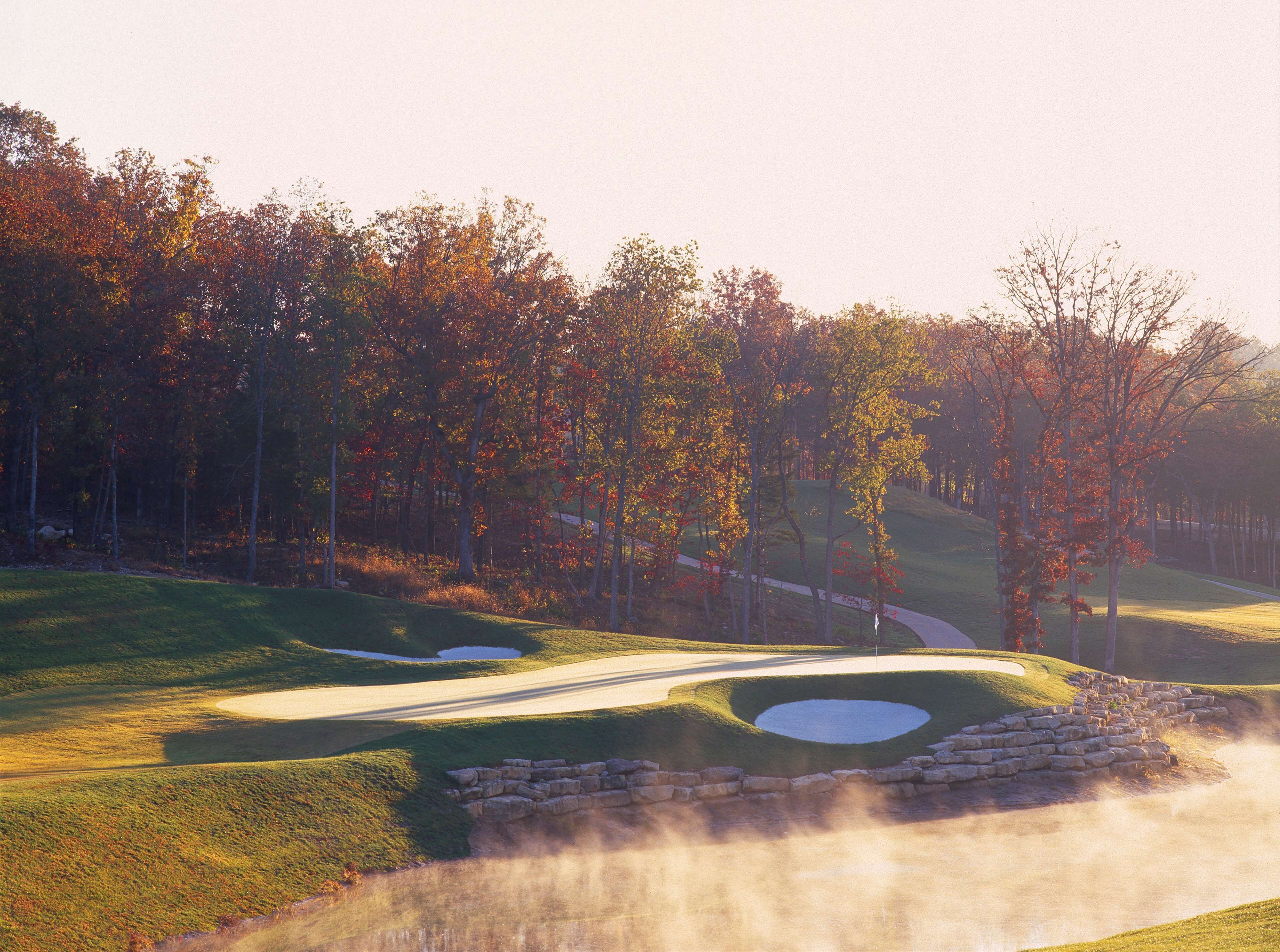 A golf course with a well-maintained fairway, sand bunkers, and a wooded area with autumn-colored trees.