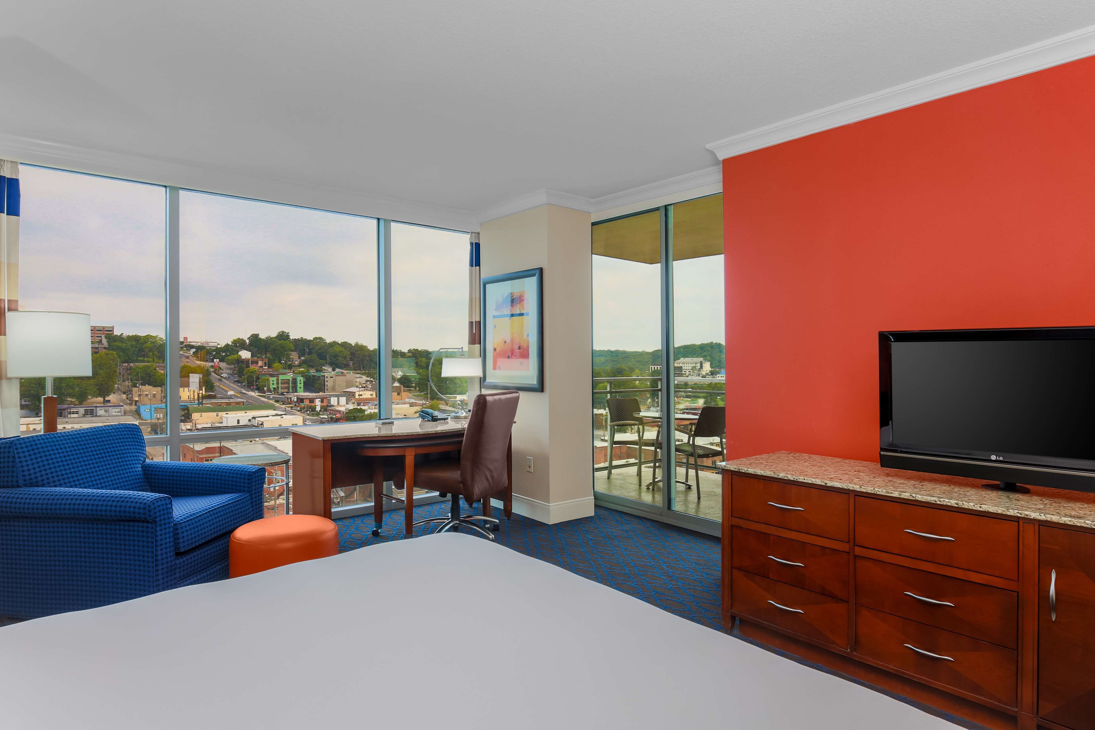 Une chambre d’hôtel avec un grand lit, un bureau, une chaise, une télévision et une vue sur la ville grâce à de grandes fenêtres.
