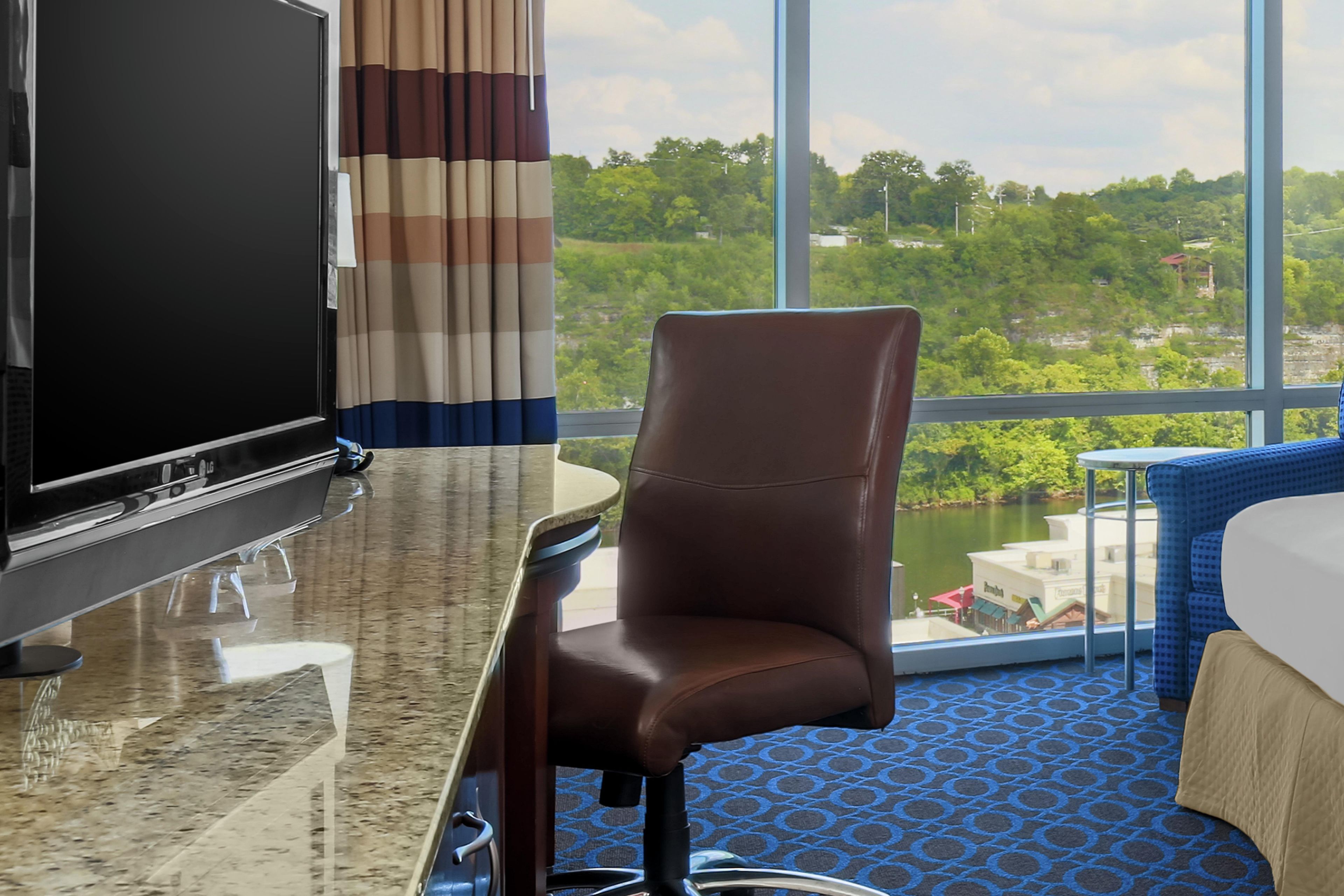 A hotel room with a large window, a flat-screen TV, a brown leather chair, a small table, and a bed with a blue patterned cover.