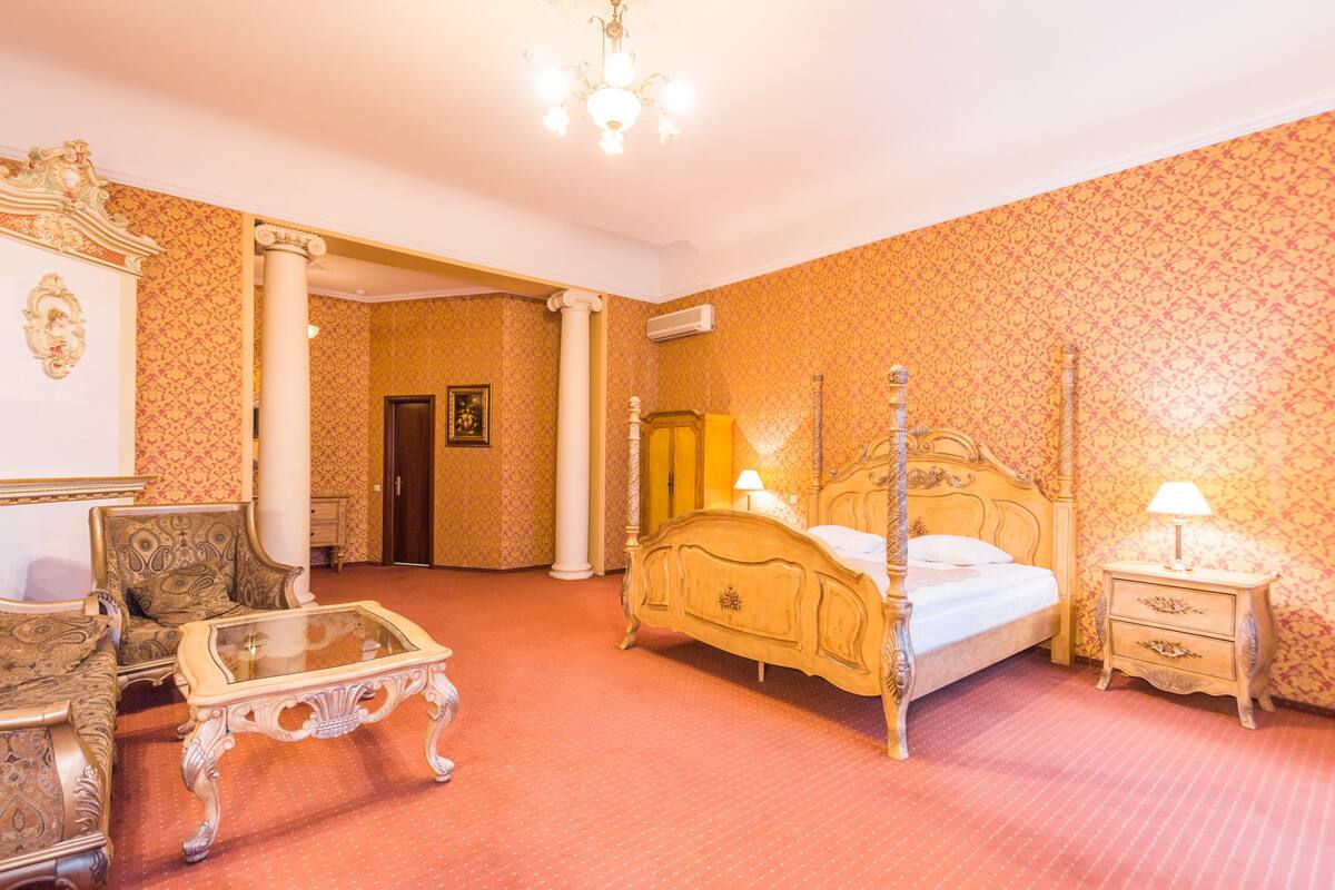 Ein Bett mit Baldachin, ein Holzkopfteil, ein Nachttisch mit Lampe und ein Couchtisch mit Glasplatte in einem Raum mit gemustertem Wandpapier und einem roten Teppich.