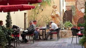 An outdoor café with tables and chairs, surrounded by greenery and a statue.