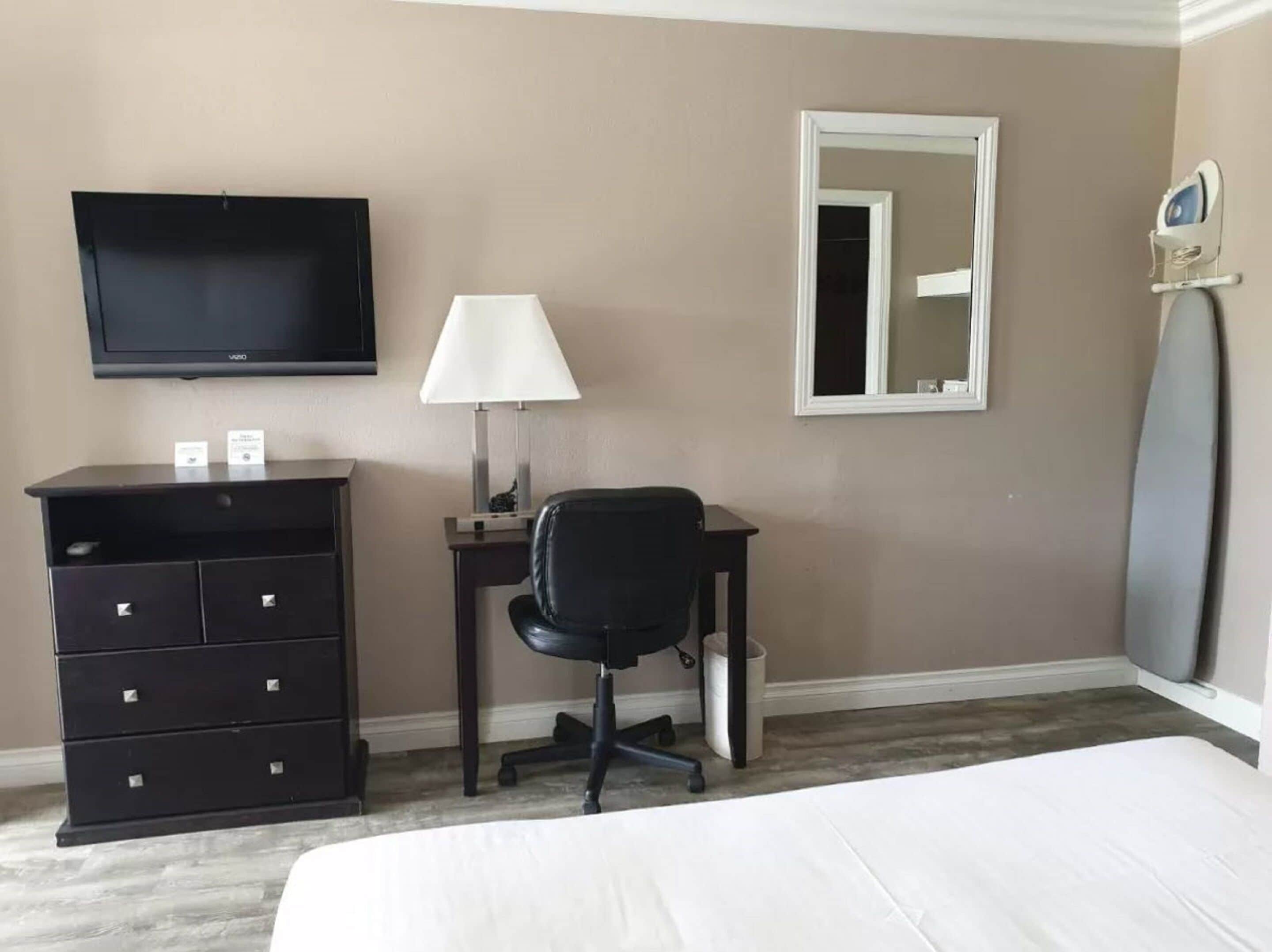 Habitación de hotel con cama, escritorio con silla, televisor, lámpara, espejo y tabla de planchar en la pared.