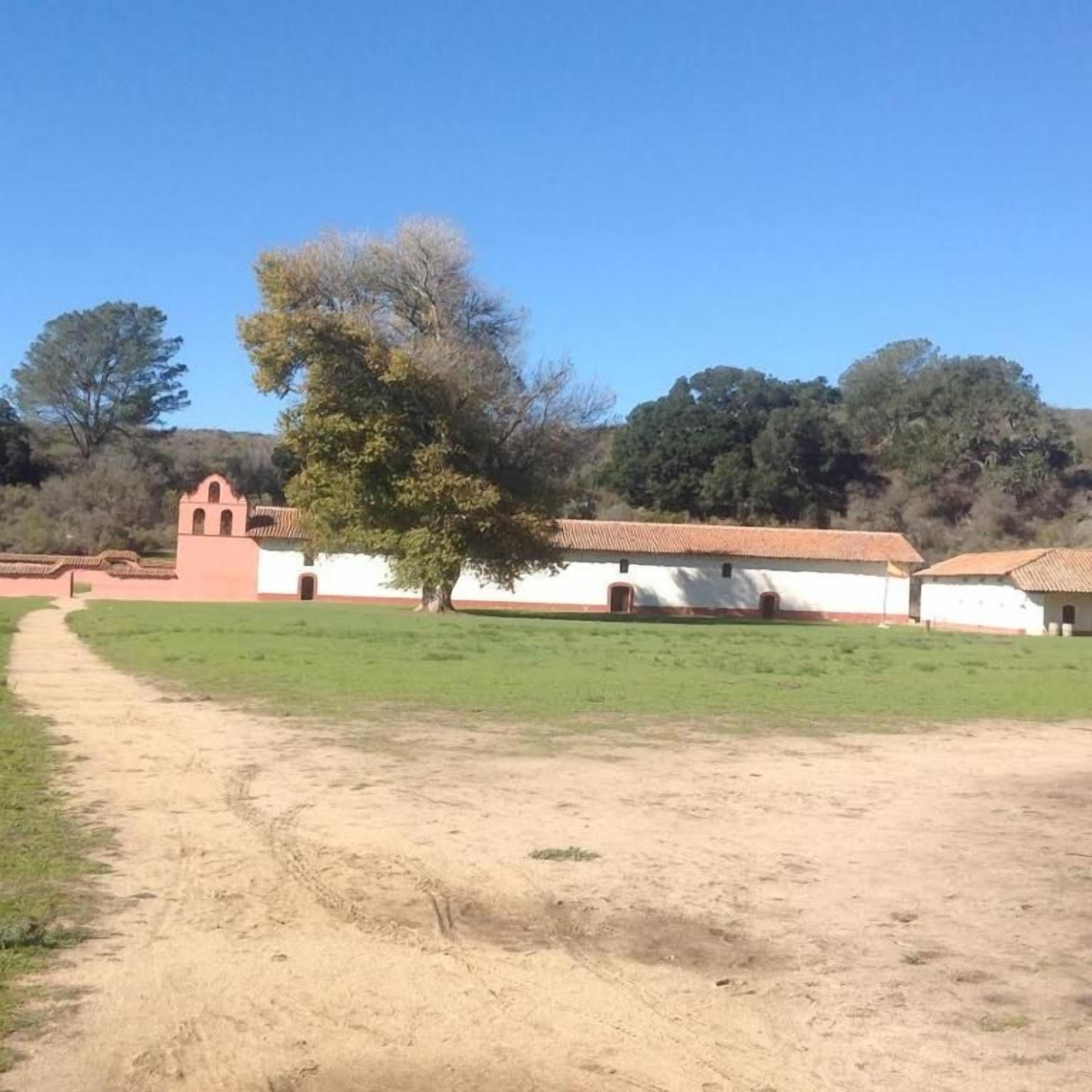 Un edificio con techo de tejas rojas y paredes blancas, un sendero de tierra que lleva a él, y un árbol en primer plano.