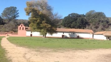 Un edificio con techo de tejas rojas y paredes blancas, un sendero de tierra que lleva a él, y un árbol en primer plano.
