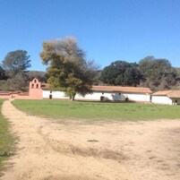 Un edificio con techo de tejas rojas y paredes blancas, un sendero de tierra que lleva a él, y un árbol en primer plano.