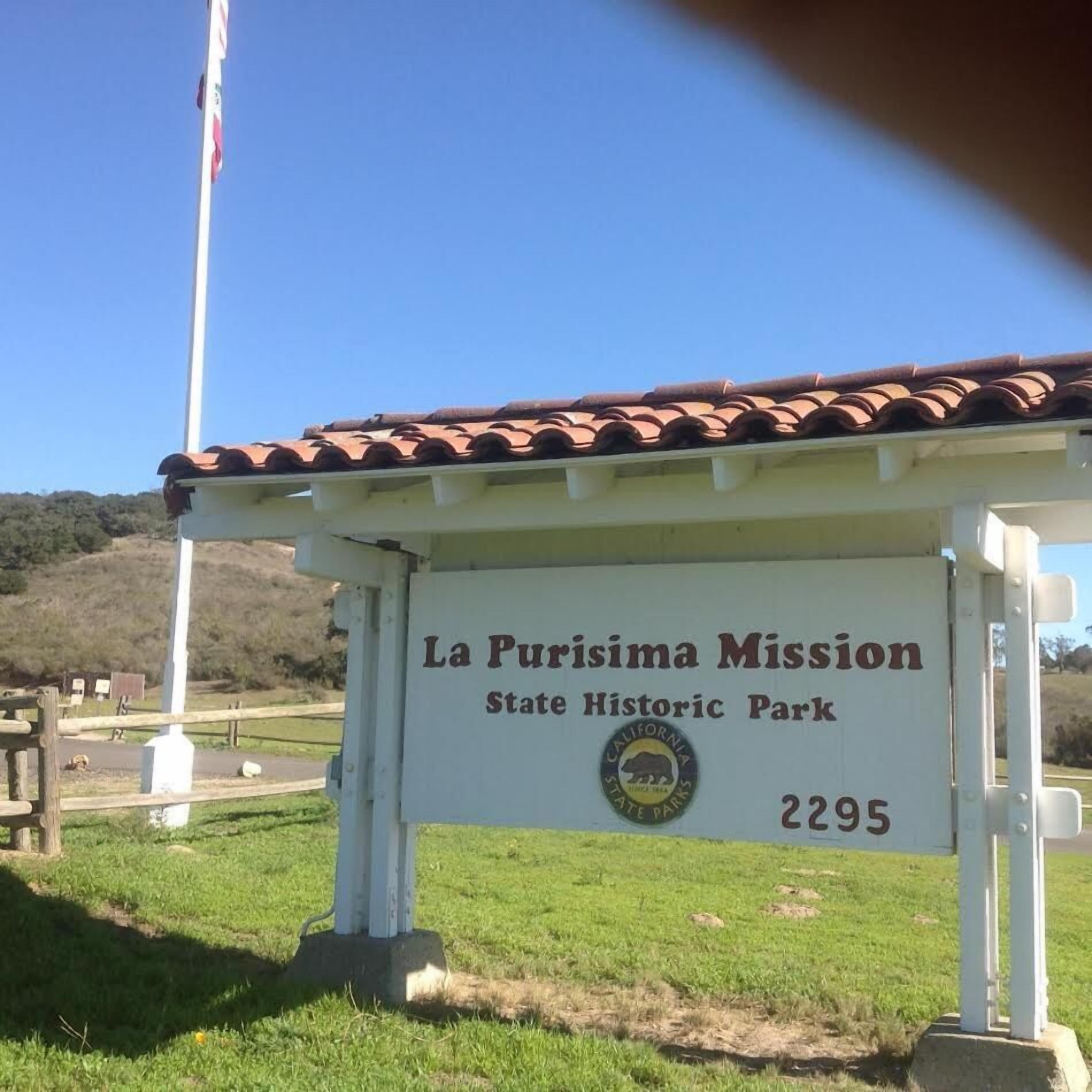A sign for La Purisima Mission State Historic Park with a clear sky and a flag in the background.