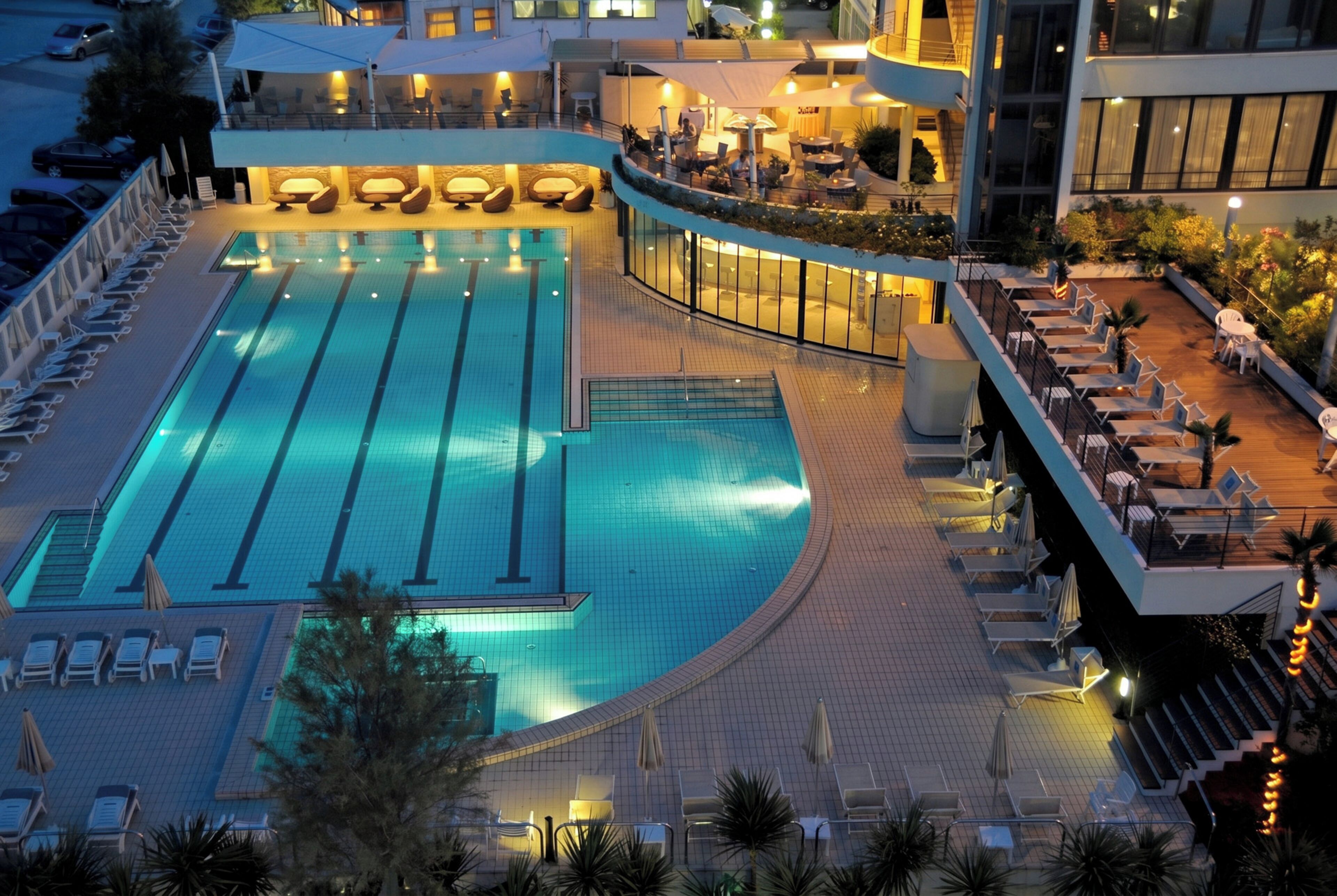Una piscina con lettini e ombrelloni, circondata da un edificio con diversi livelli e zone salotto all'aperto.