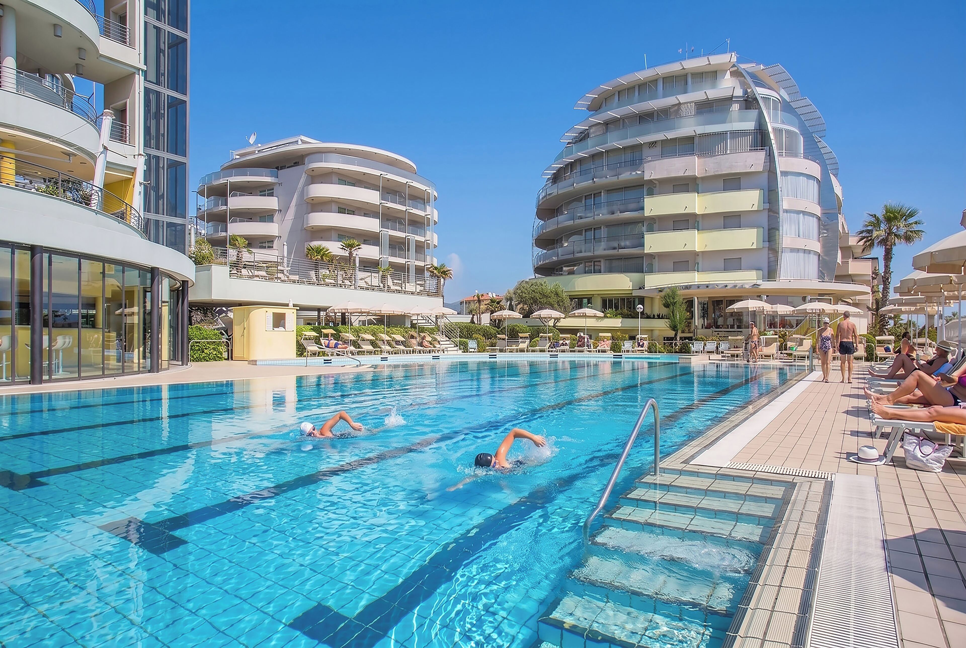 A swimming pool with multiple lanes, surrounded by modern buildings with balconies and outdoor seating.
