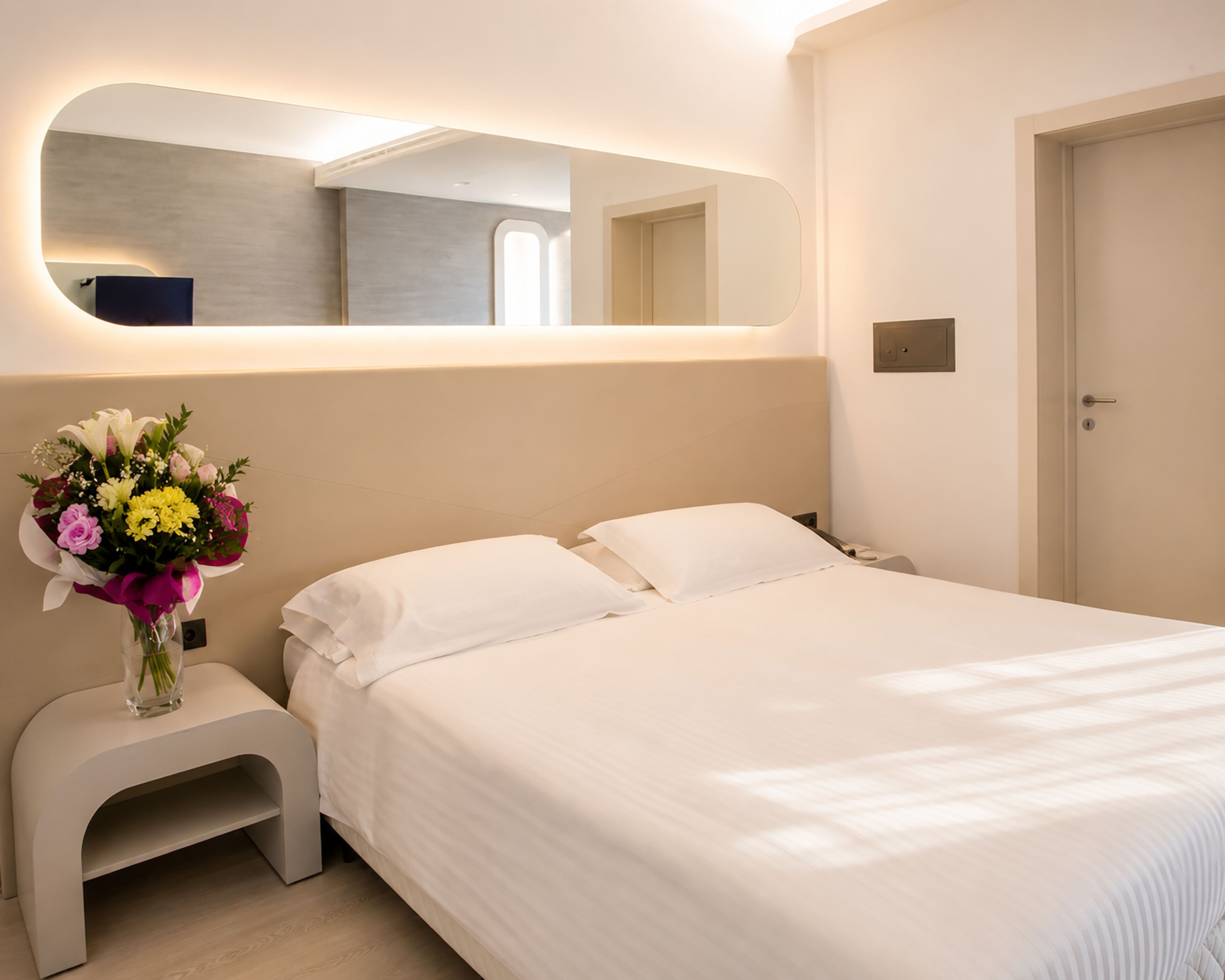 A hotel room with a bed, a nightstand, a vase of flowers, a mirror, and a door.