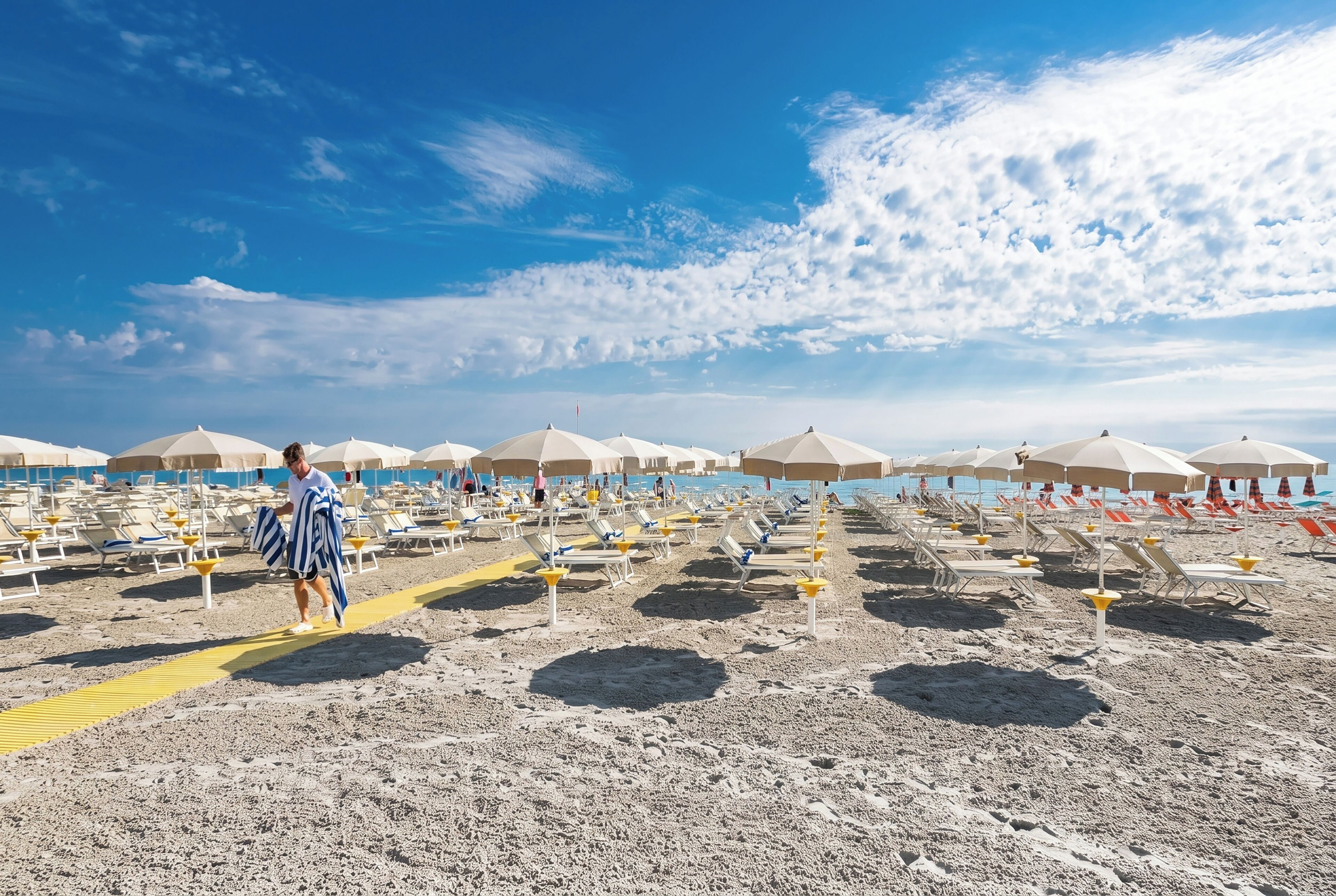 Een strand met rijen ligbedden en parasols, een heldere lucht en iemand die loopt.