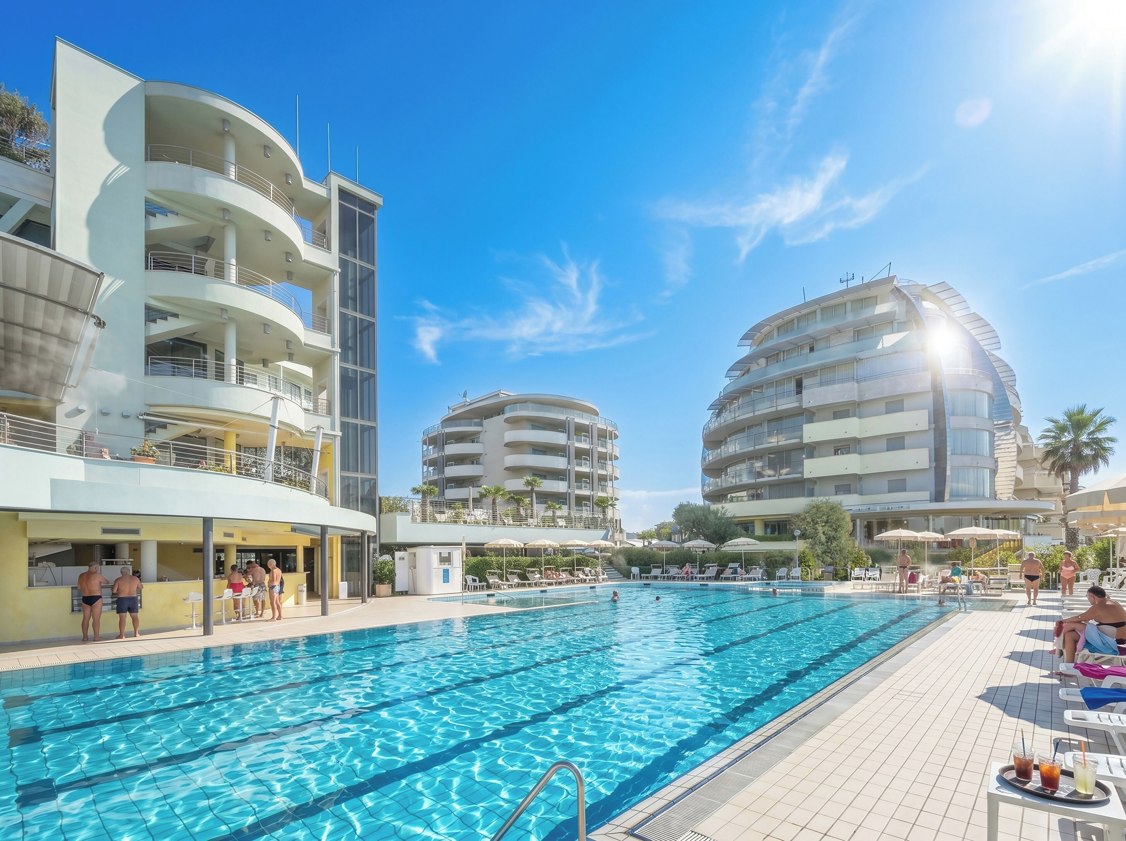 A swimming pool with a sunlit sky, surrounded by modern buildings with balconies and outdoor seating.