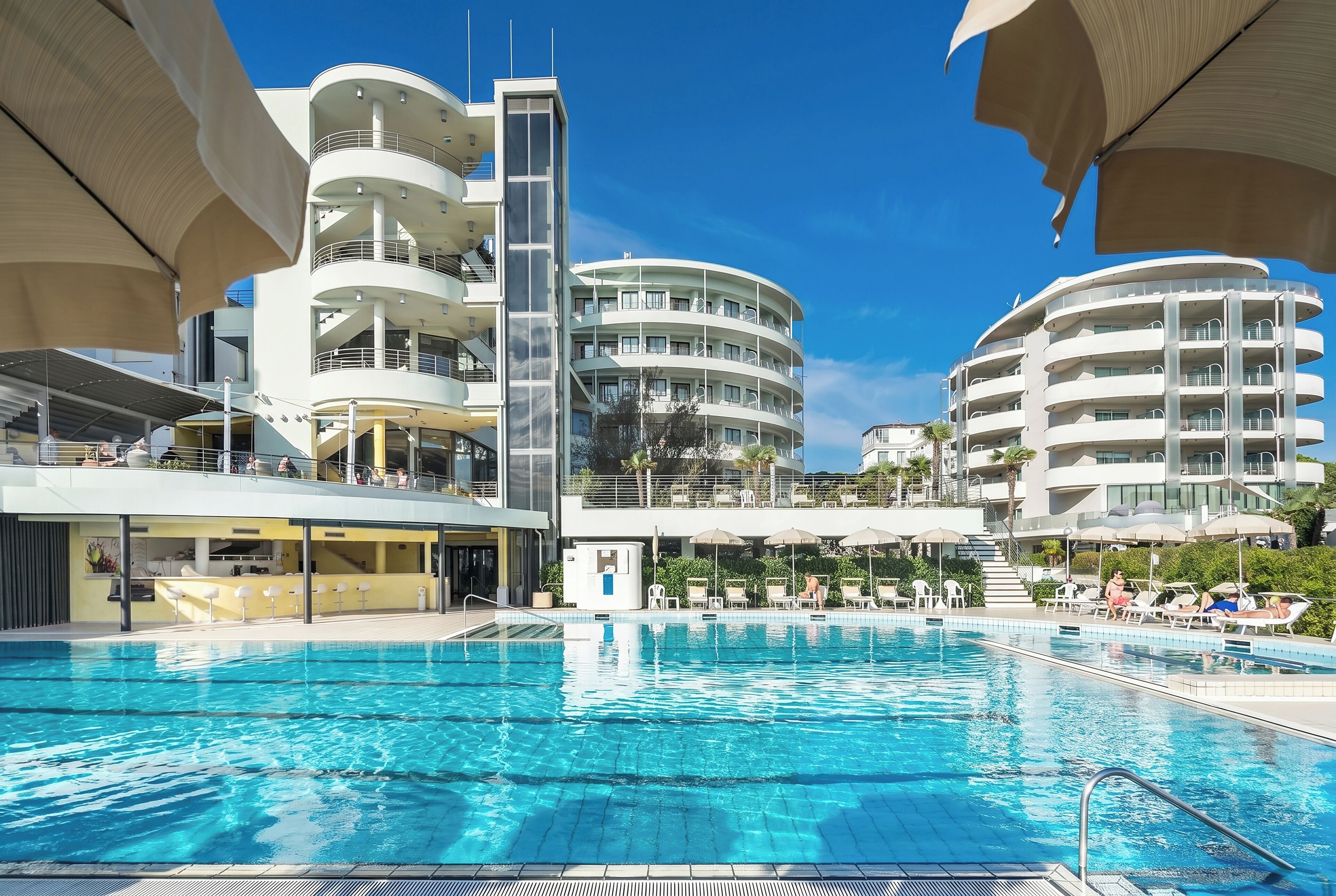 A swimming pool with a modern building in the background, featuring multiple balconies and a clear blue sky.