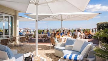 Een terras buiten met tafels, stoelen, parasols en uitzicht op zee.