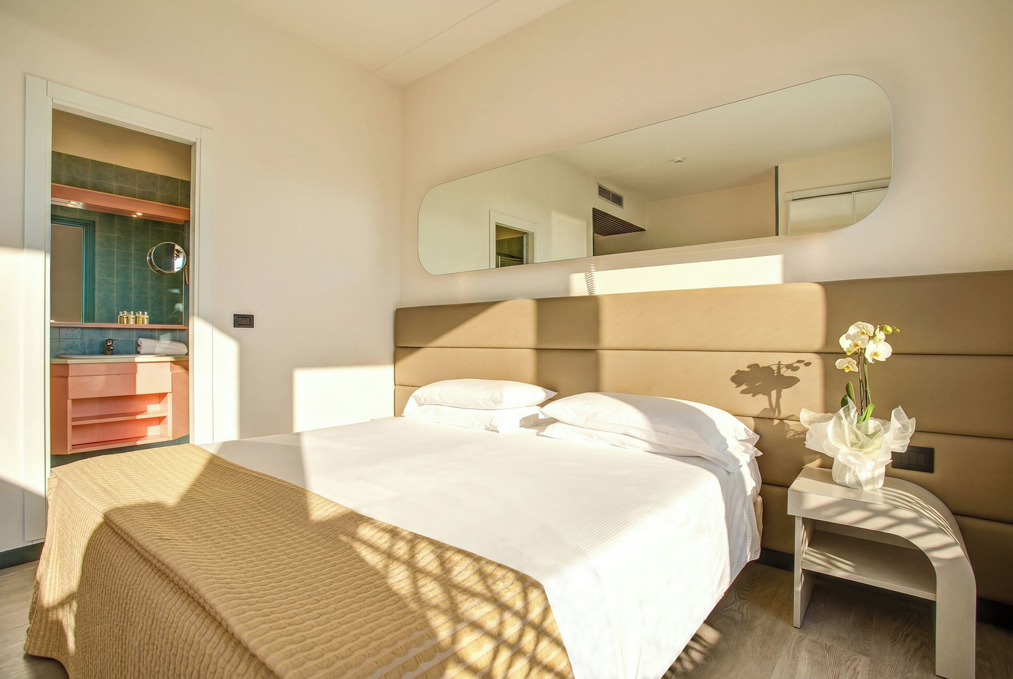 Habitación de hotel con cama, mesita de noche con un florero, un espejo y baño con lavamanos y espejo.