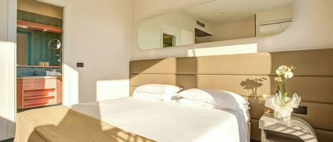 Habitación de hotel con cama, mesita de noche con un florero, un espejo y baño con lavamanos y espejo.