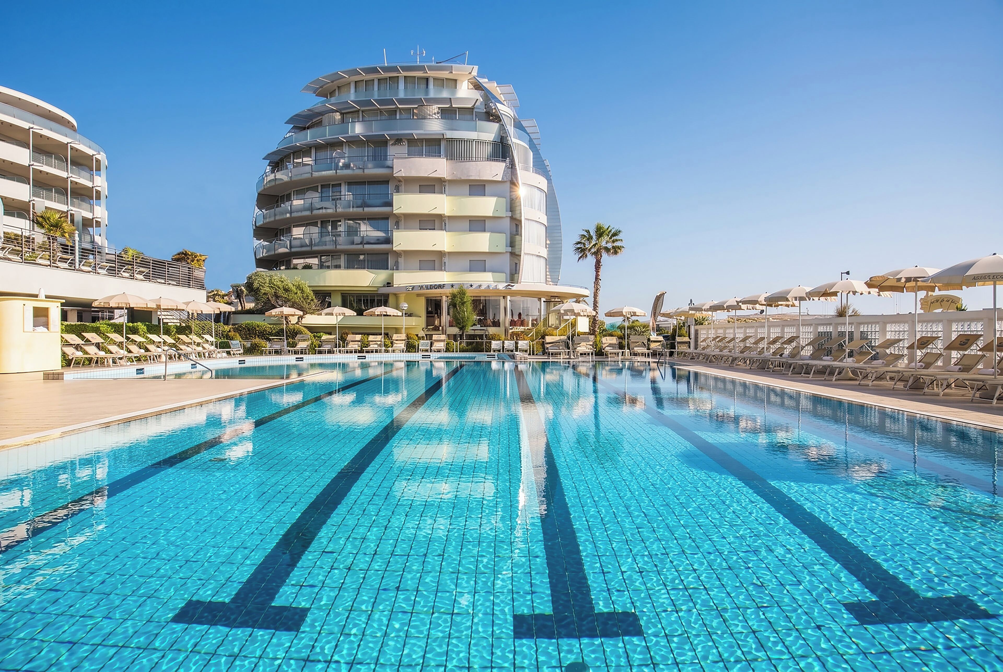 Een modern hotel met een groot zwembad, meerdere balkons en zonneschermen.