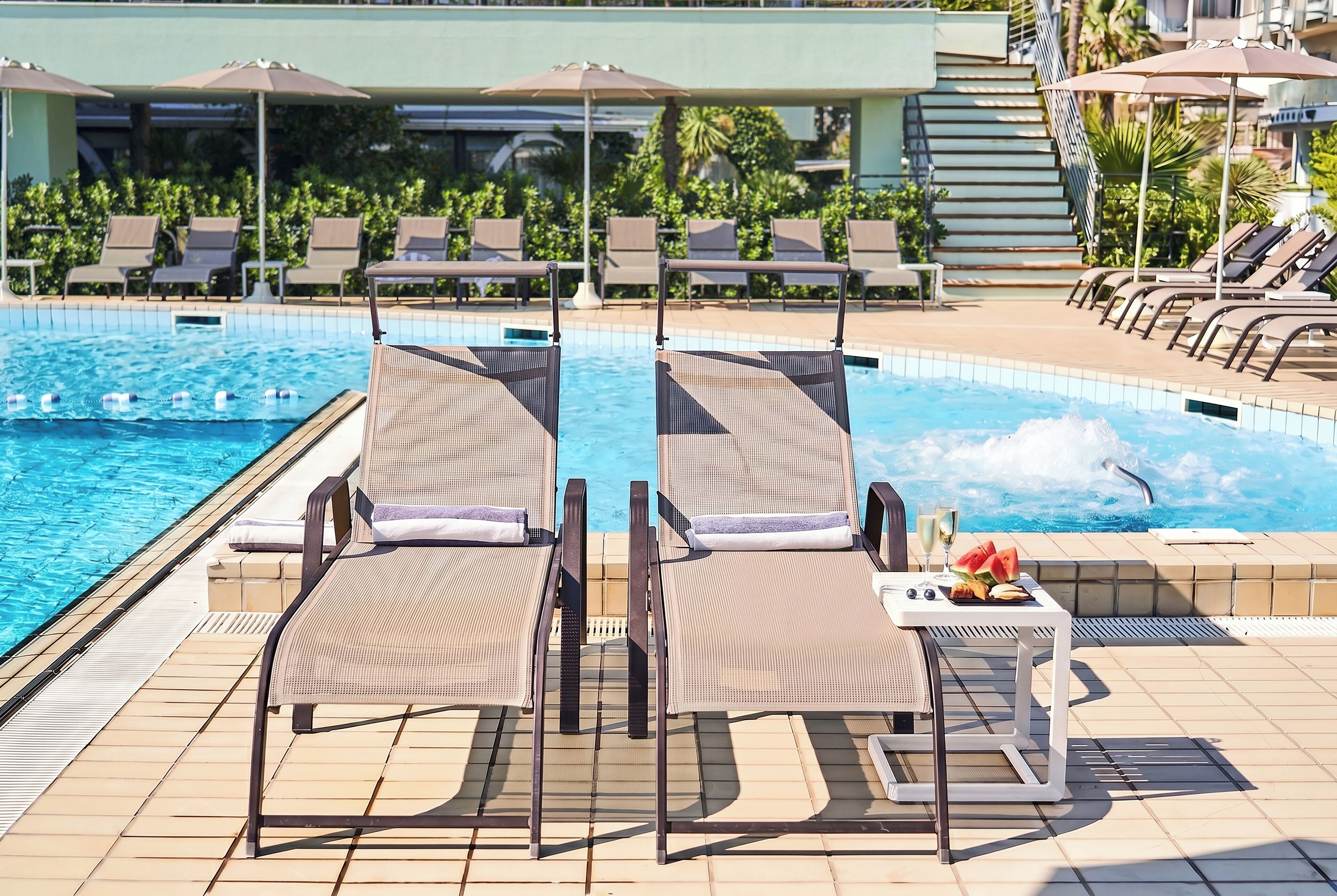 Zona de piscina con sillones de descanso y sombrillas, un suelo de baldosas y una escalera que lleva a un nivel superior.