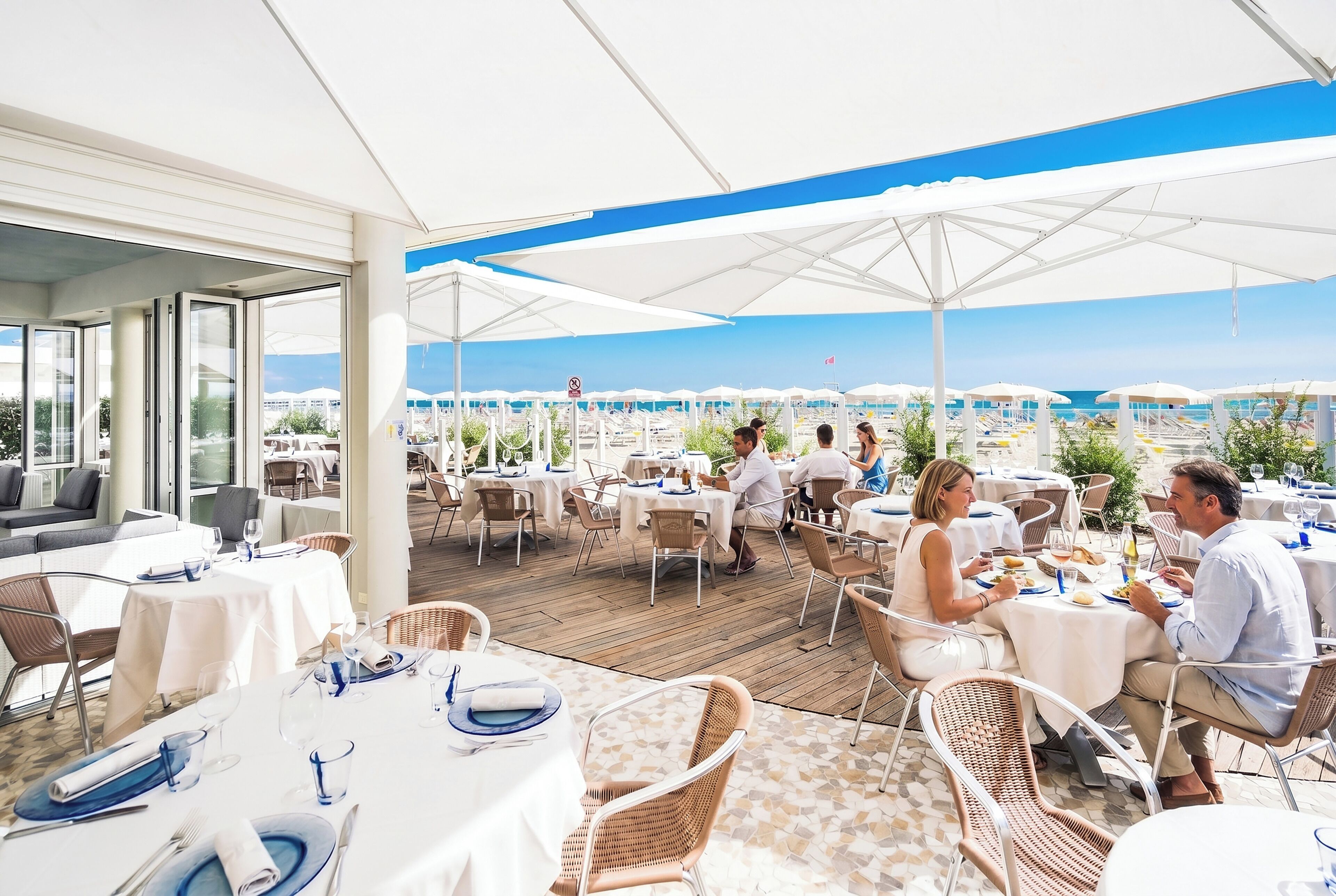 An outdoor dining area with tables and chairs, a view of the beach, and umbrellas providing shade.
