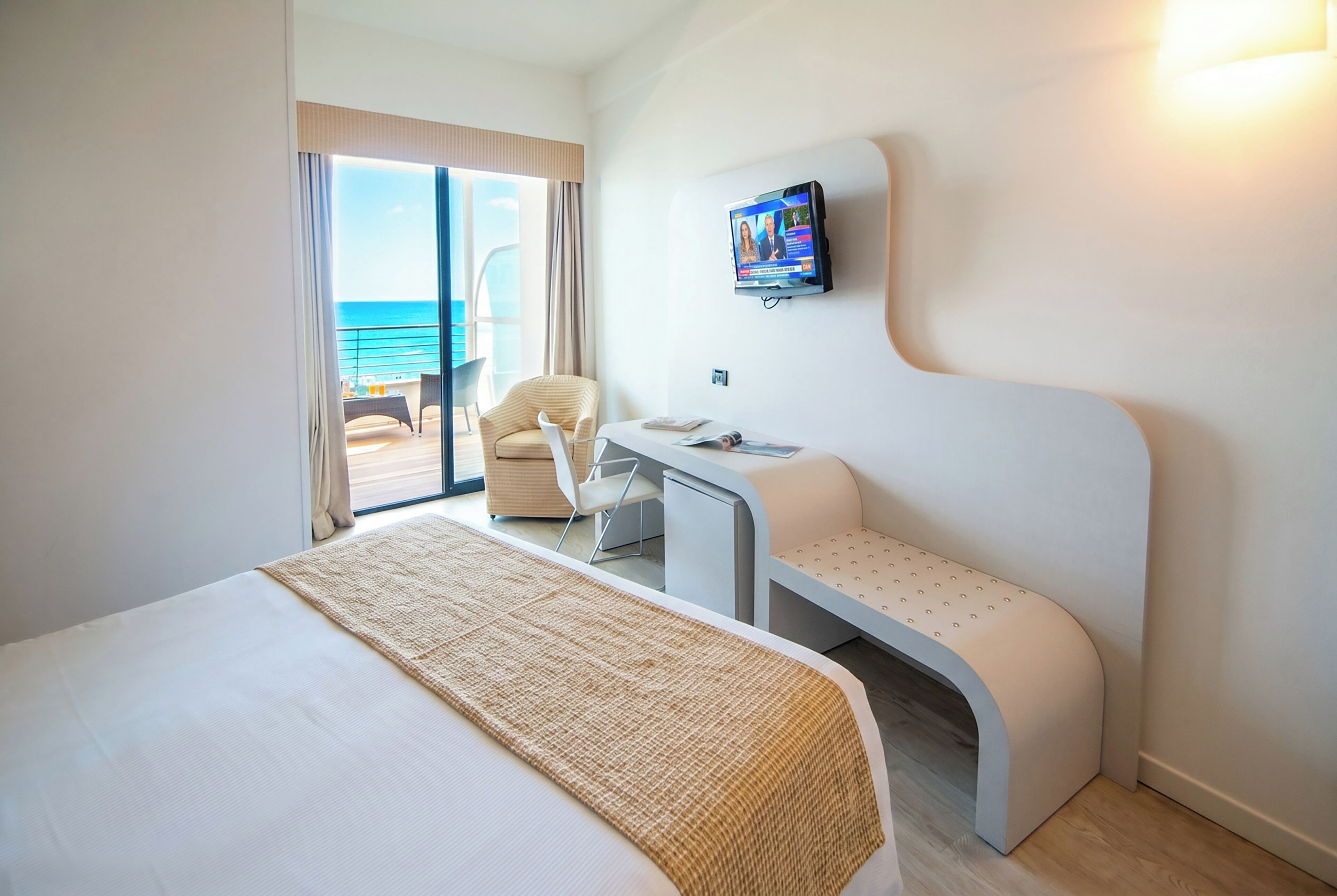 A hotel room with a bed, a desk, a chair, a television, and a view of the ocean.