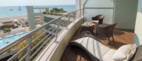 A balcony with wicker furniture, a glass partition, and a view of the sea and pool.