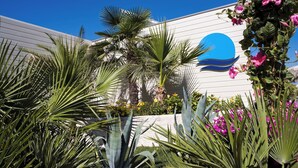 A building with a white exterior and a blue circular logo, surrounded by various tropical plants and flowers. - Hotel Le Palme (Cervia)