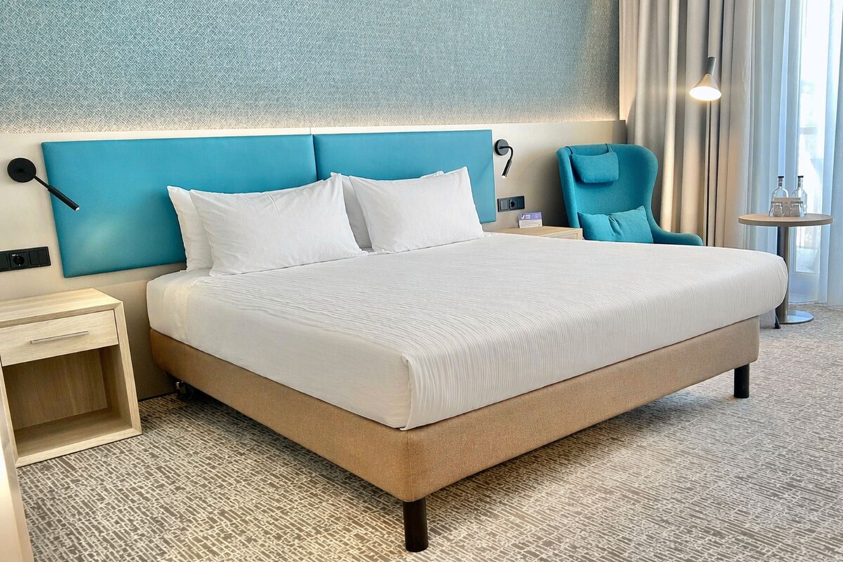 A hotel room with a large bed, a bedside table, a chair, a small table, and a window with curtains.