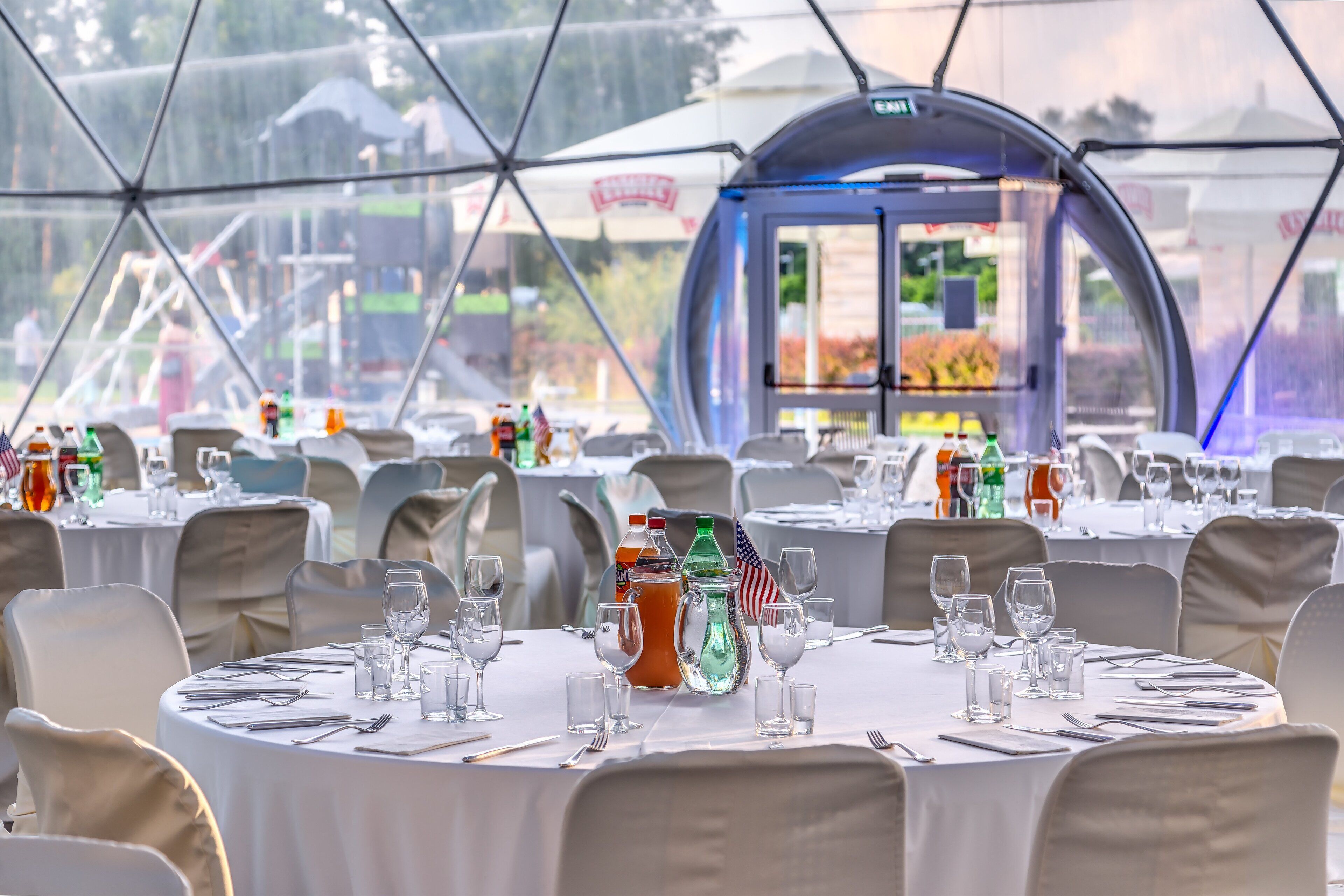 A tented dining area with round tables set for service, featuring clear glassware and bottled beverages.