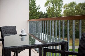 A glass-top table with a white cup on a wicker chair, a metal railing, and a view of trees. - Holiday Inn Resort Warszawa Józefów**** by IHG (Jozefow)