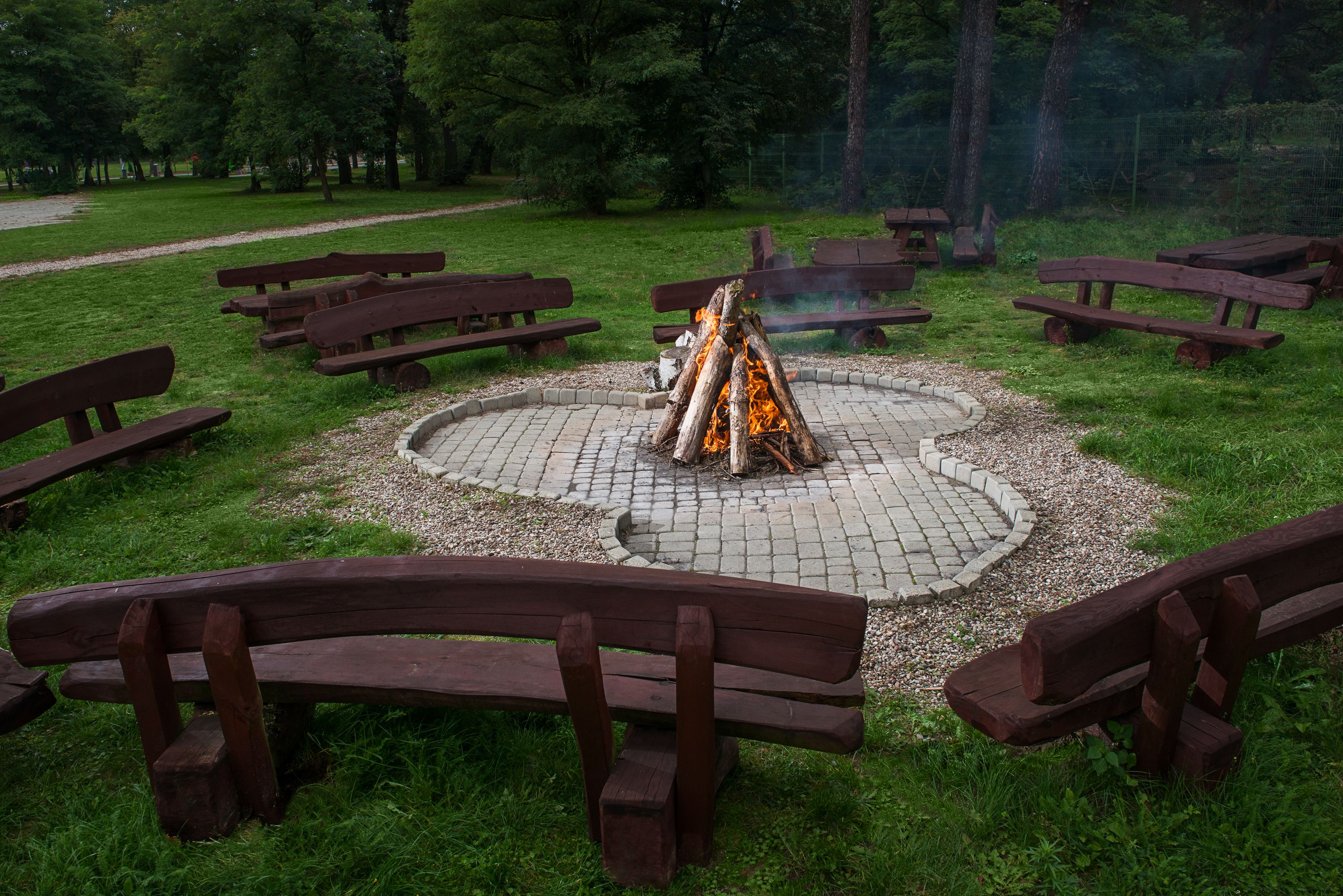 A circular fire pit surrounded by wooden benches in a park setting.