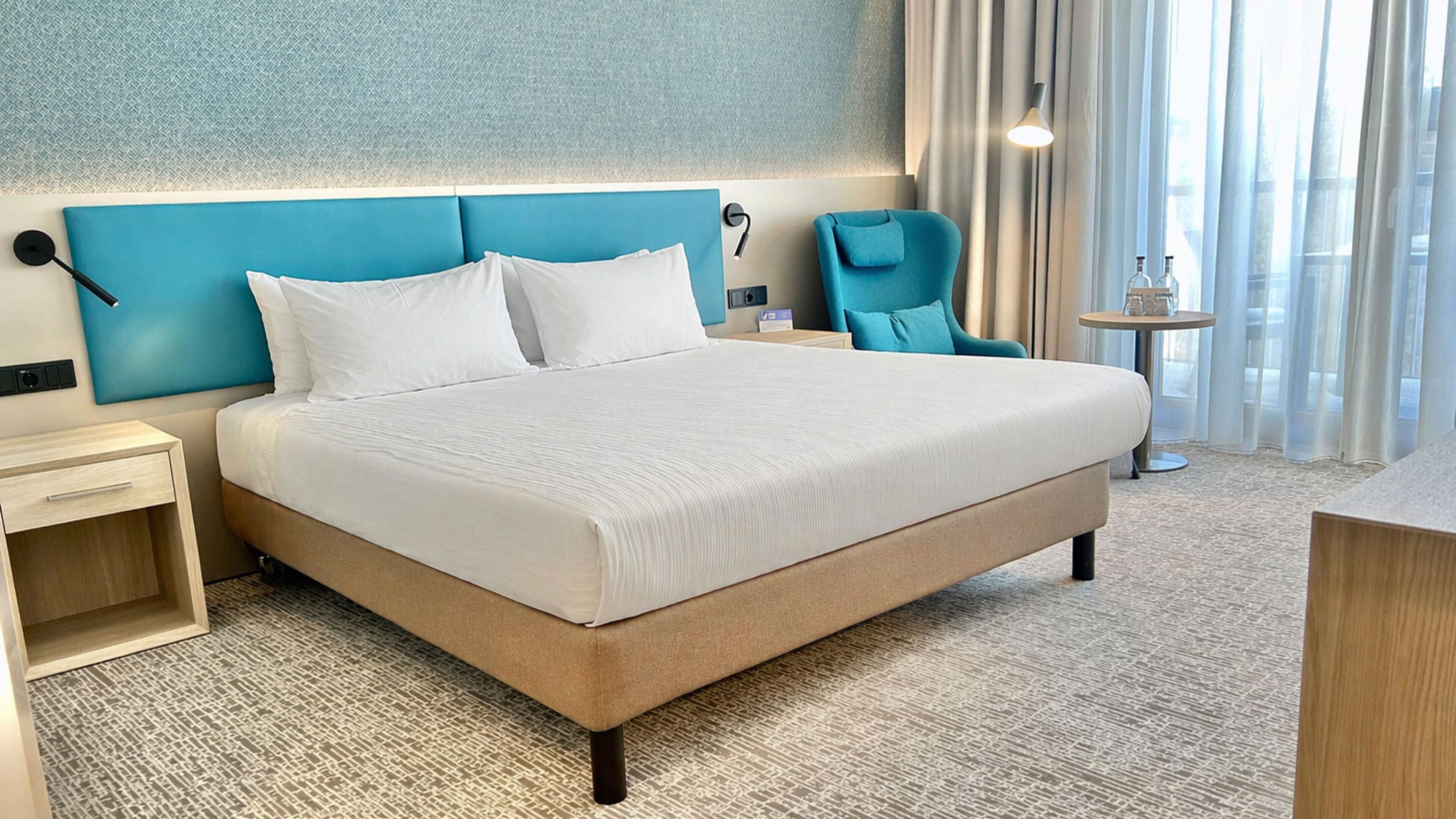 A hotel room with a large bed, a bedside table, a chair, a small table, and a window with curtains.