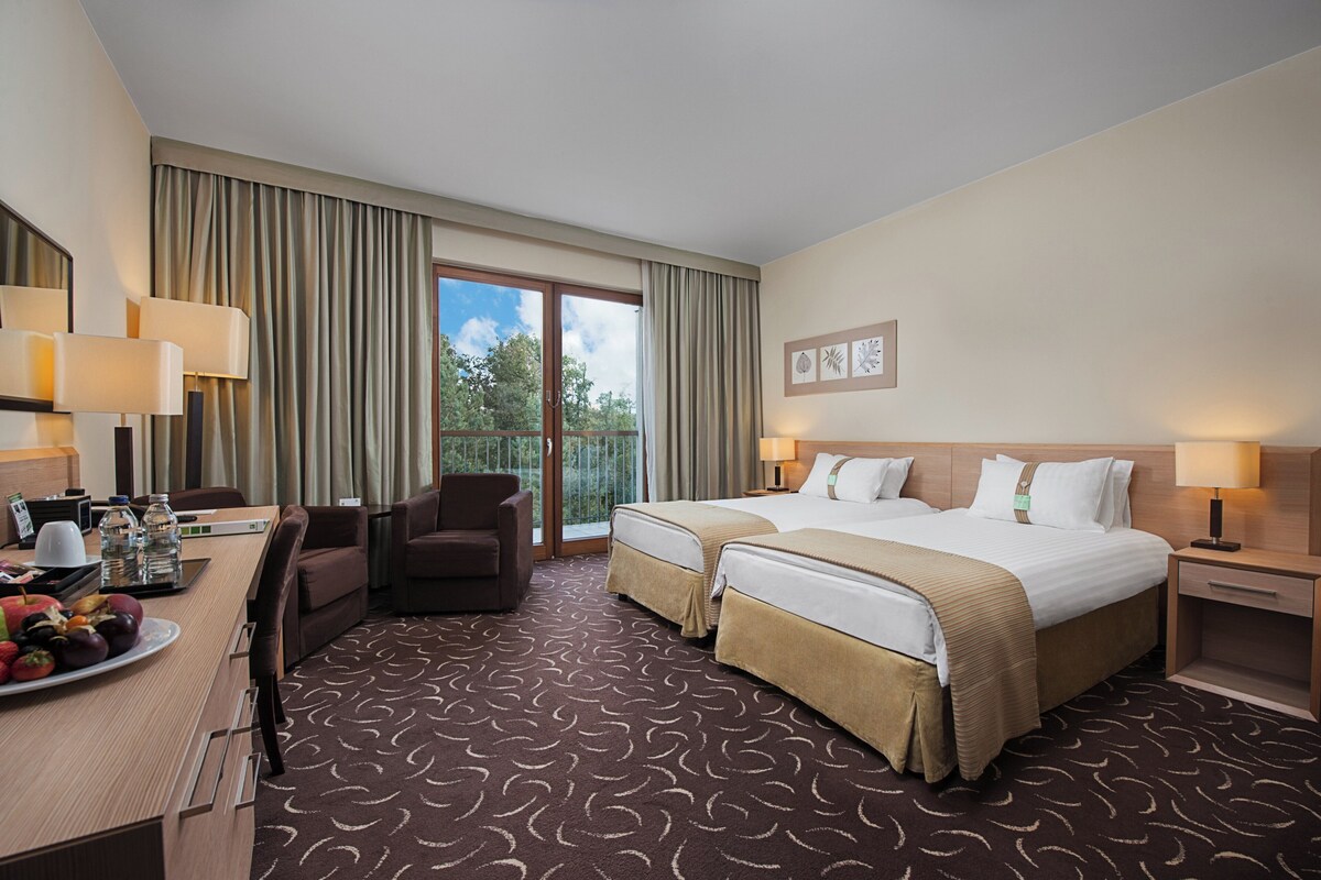 A hotel room with two beds, a desk with a chair, a small table with fruit, and a view of trees through the window.