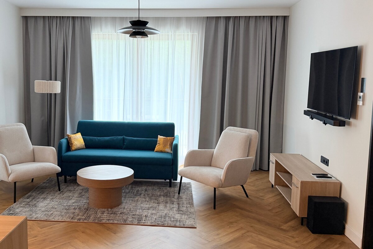 A modern living room with a teal sofa, two armchairs, a coffee table, and a TV mounted on the wall.