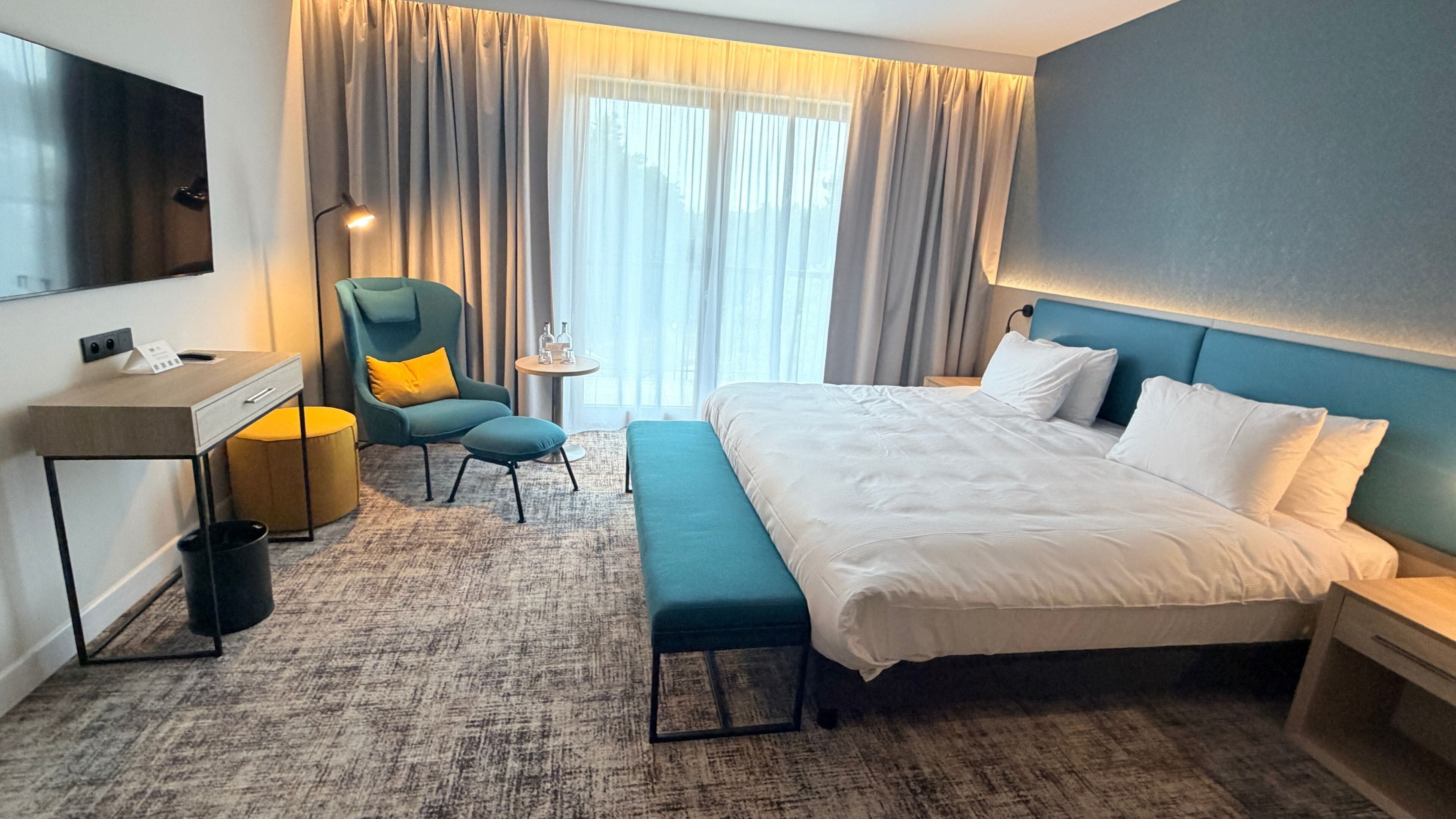 A hotel room with a large bed, a desk, a chair, a small table, and a trash bin.
